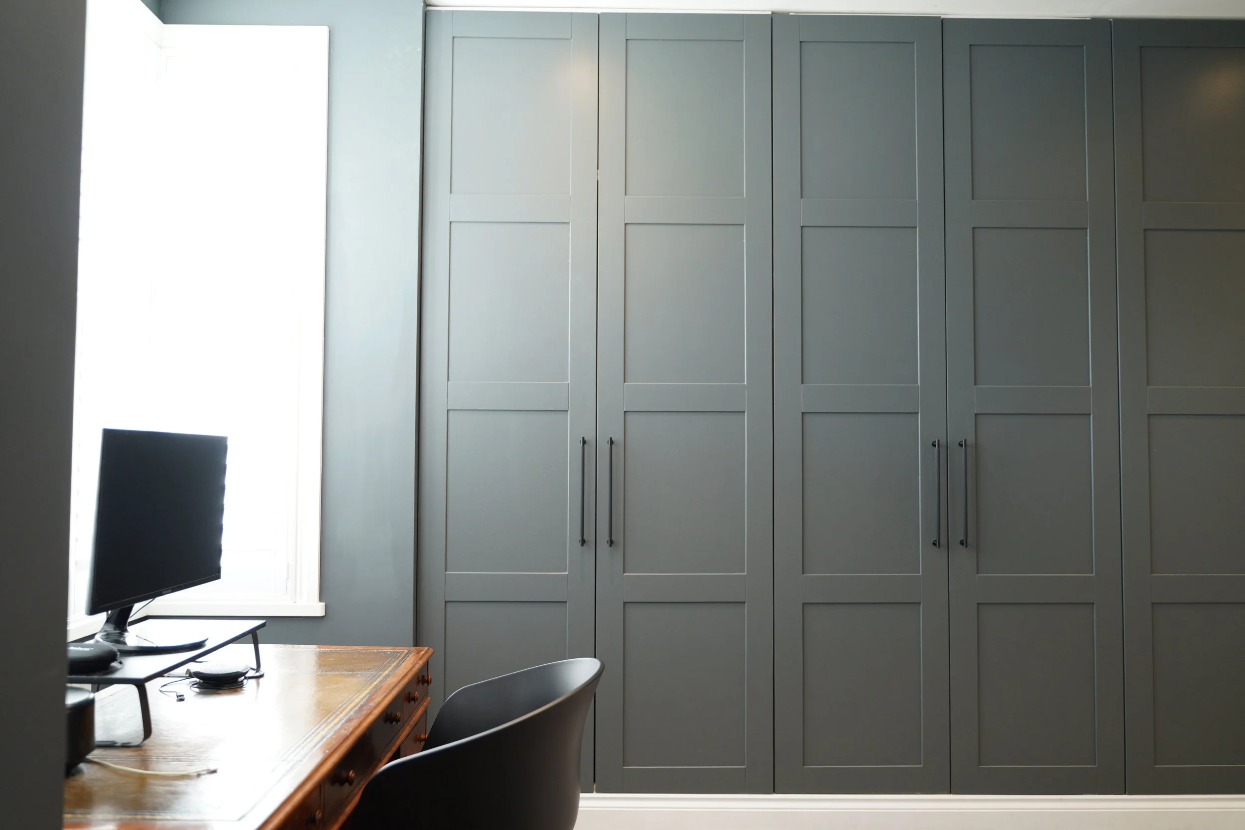 A home office with a large gray built-in cabinet, a wooden desk with a computer monitor, a black chair, and a window with bright natural light