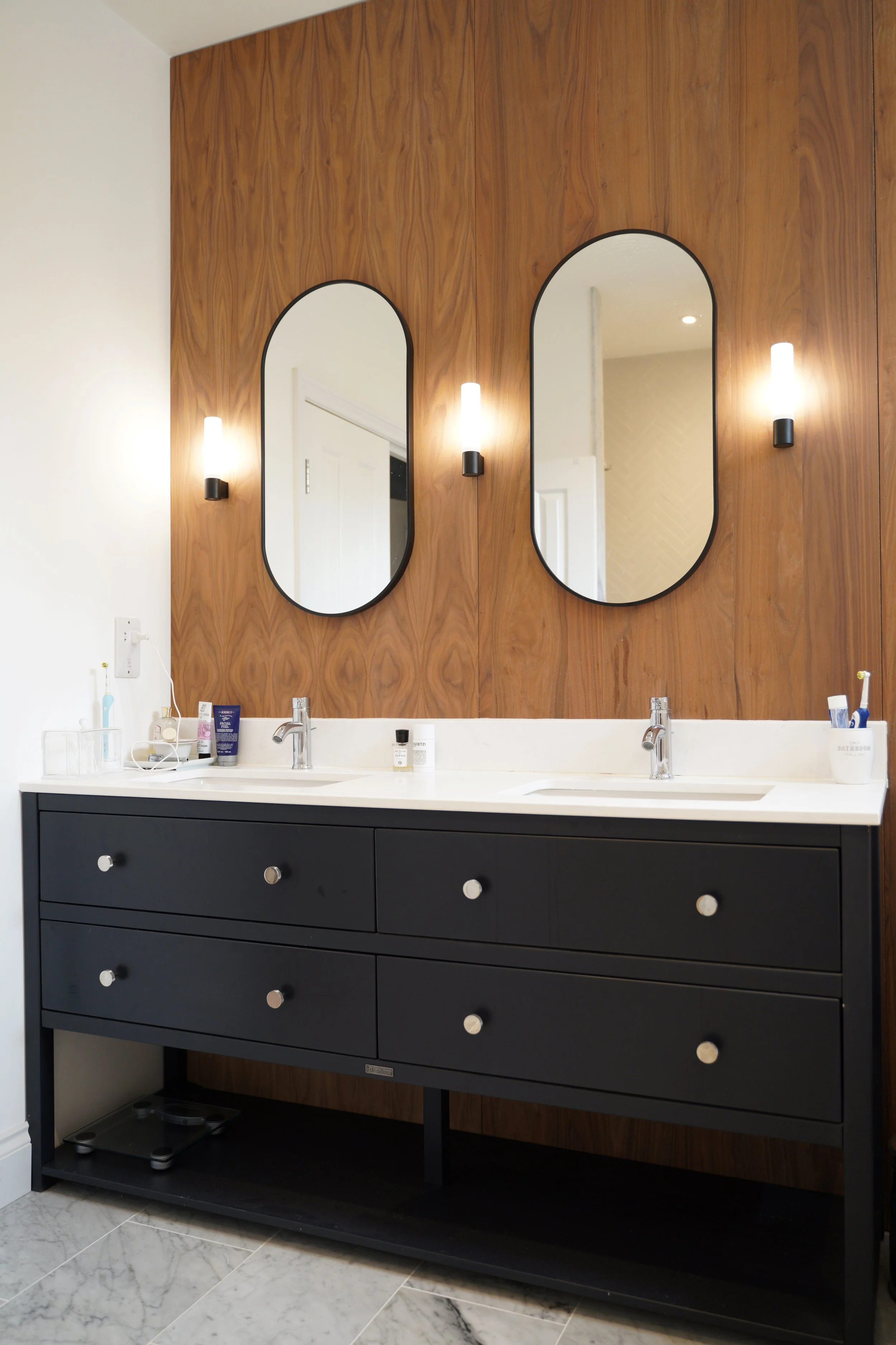A modern bathroom vanity with two sinks, black cabinet with silver knobs, and two oval mirrors on a wooden wall. There are two wall sconces with white bulbs between the mirrors, and bathroom essentials like soap, toothbrushes, and skincare products o