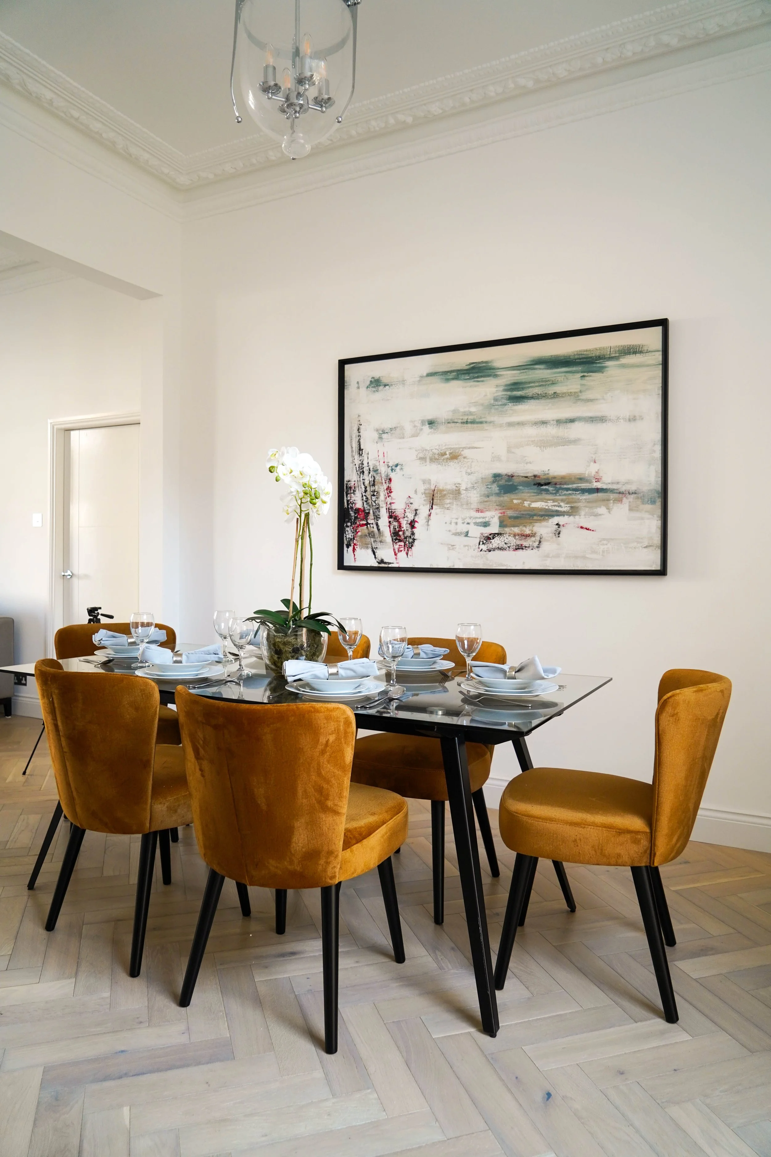 A dining room with a glass table set with white dinnerware, napkins, and wine glasses. Golden-yellow upholstered chairs surround the table. A flowering plant in a vase is at the center. An abstract painting hangs on the white wall. A ceiling light fi