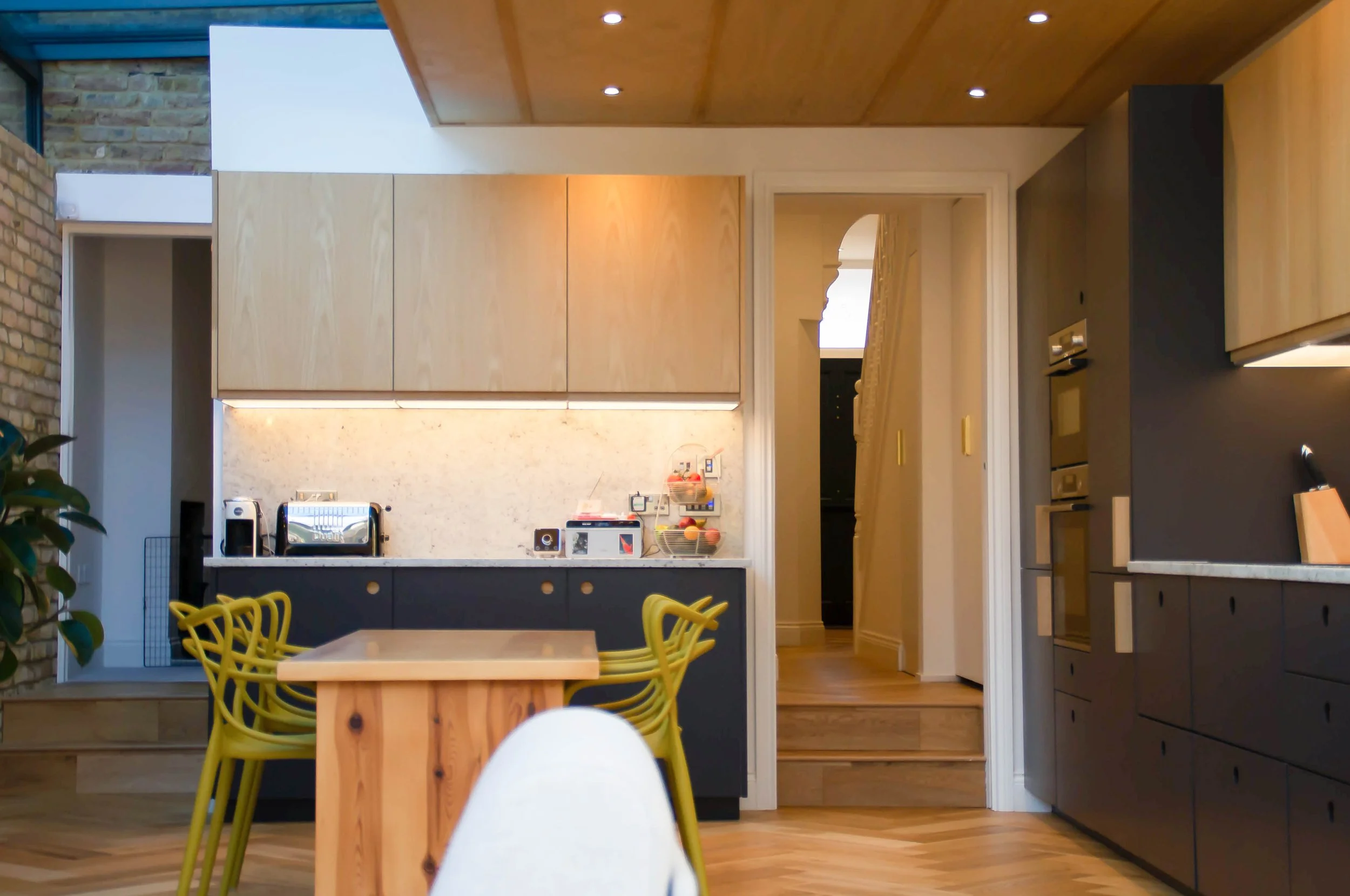 Modern kitchen with wooden cabinets, black lower cabinets, a wooden dining table with yellow chairs, and a hallway leading to another room.