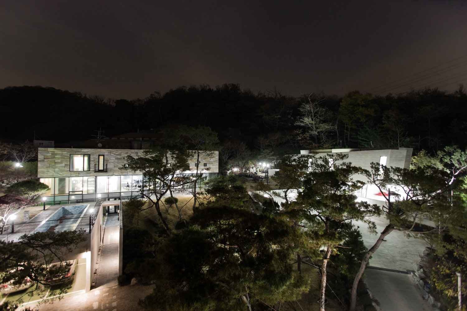 Nighttime view of modern buildings with illuminated windows, surrounded by trees, with a hill and dark sky in the background.