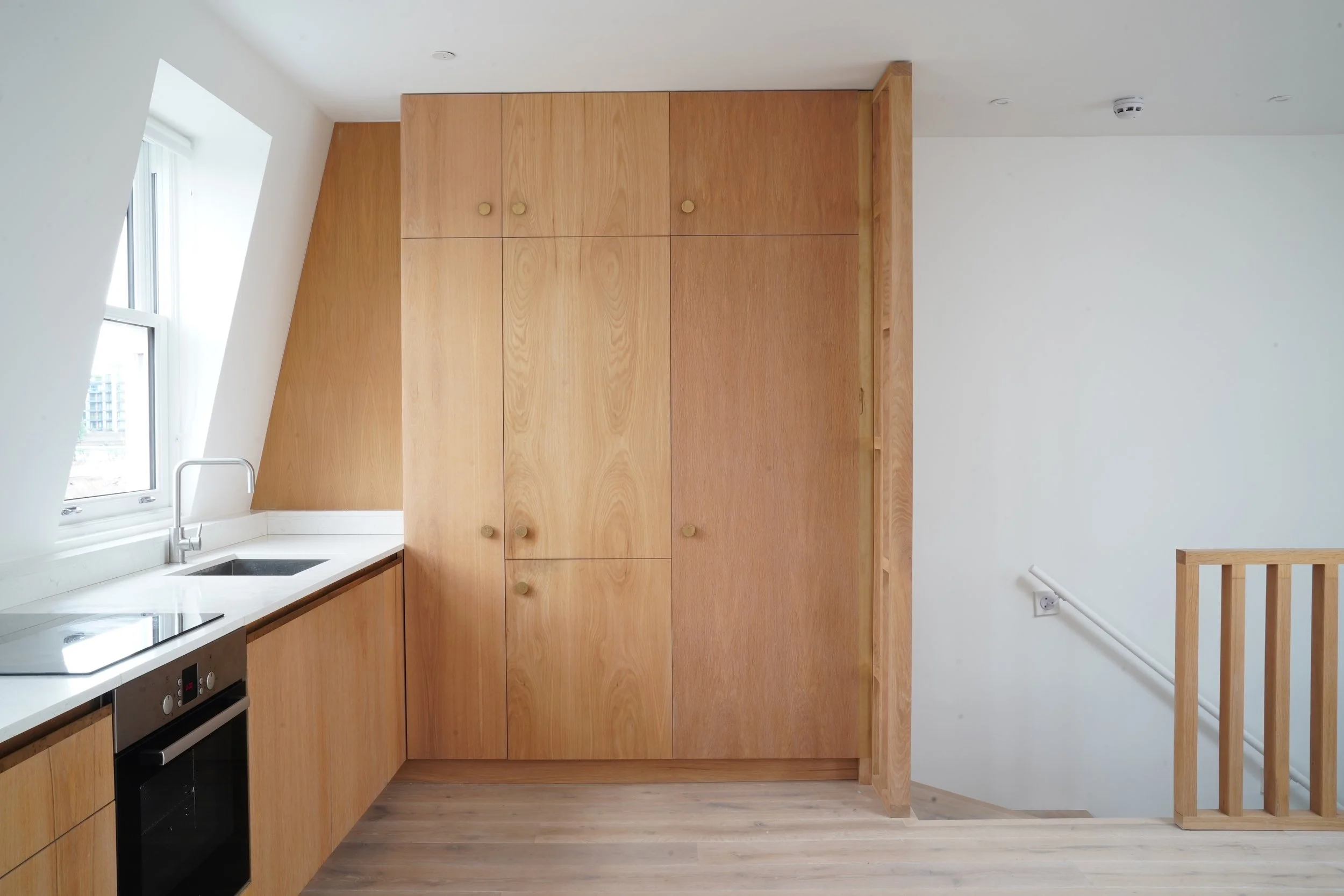 Interior of a modern kitchen with white walls and wooden cabinetry, a window over the sink, and a staircase with a wooden railing.
