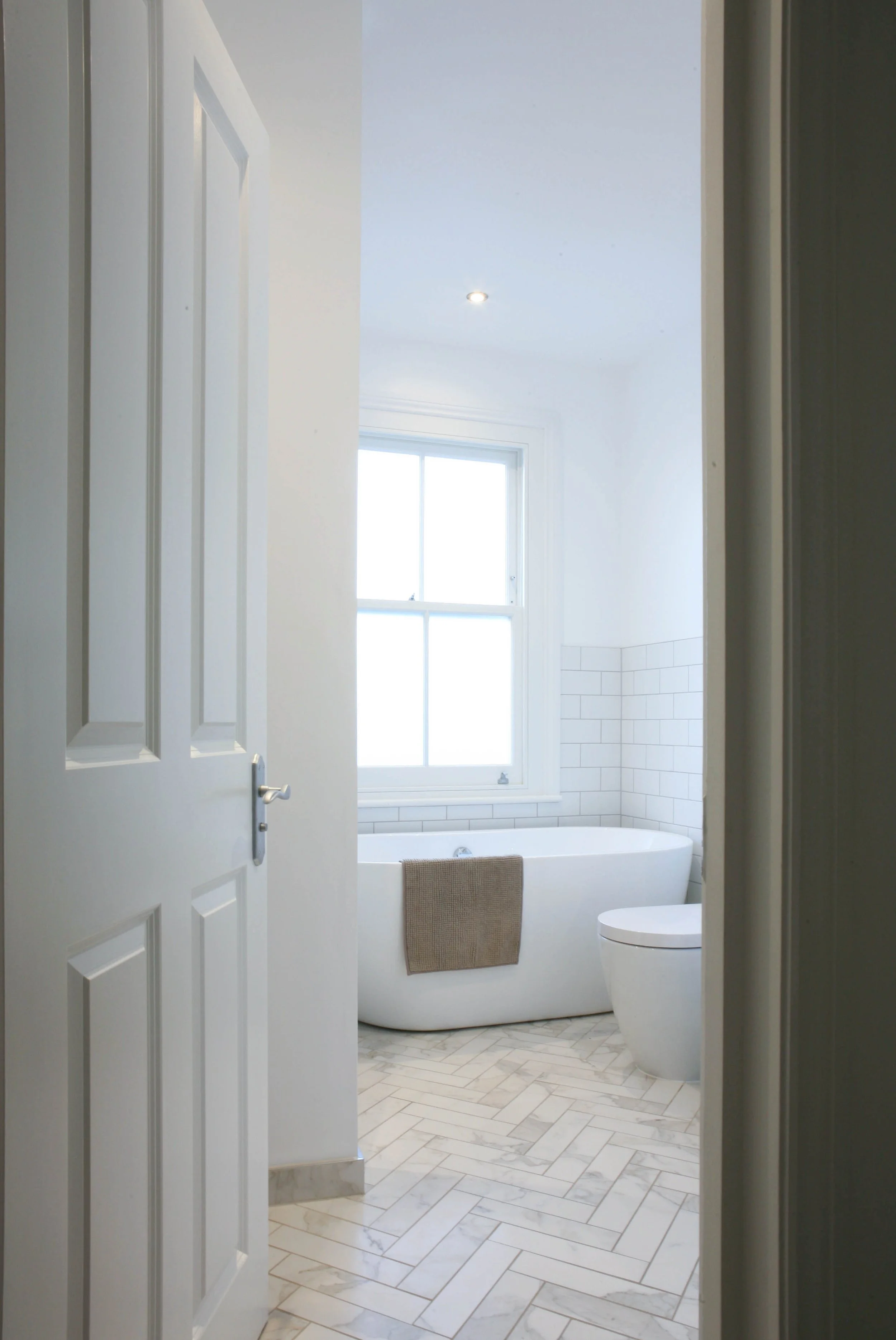 View into a modern, bright bathroom with a bathtub, toilet, large window, white tile walls, and herringbone marble floor.