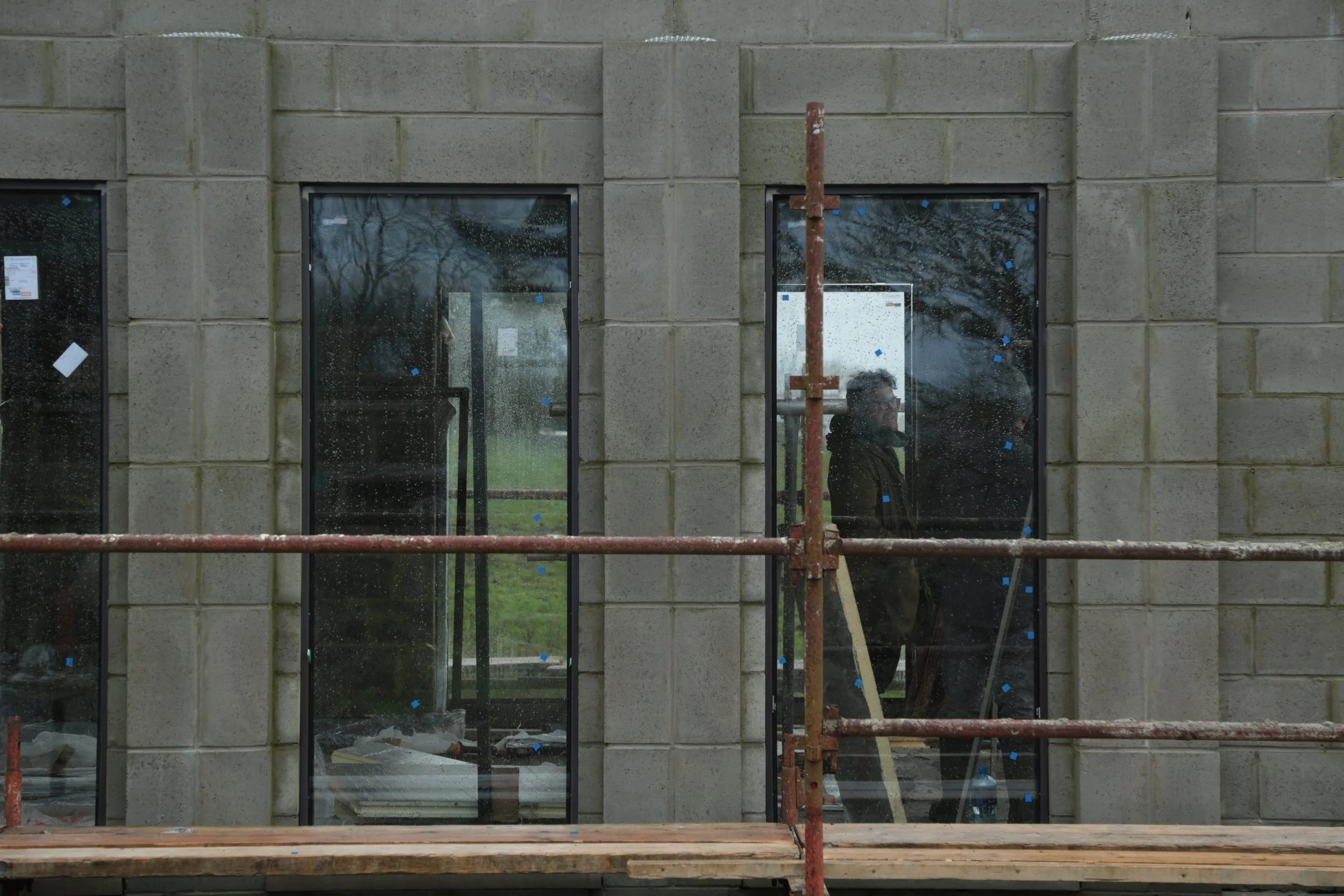 A construction worker in black jacket and glasses standing behind scaffolding outside a building with large glass windows, some of which have blue stickers, during rainy weather.