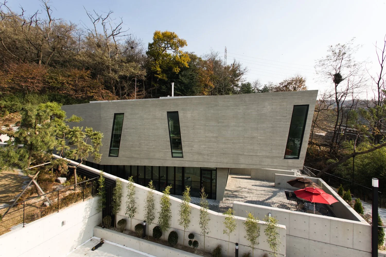 Modern concrete building with angled windows, surrounded by trees and outdoor seating with umbrellas.