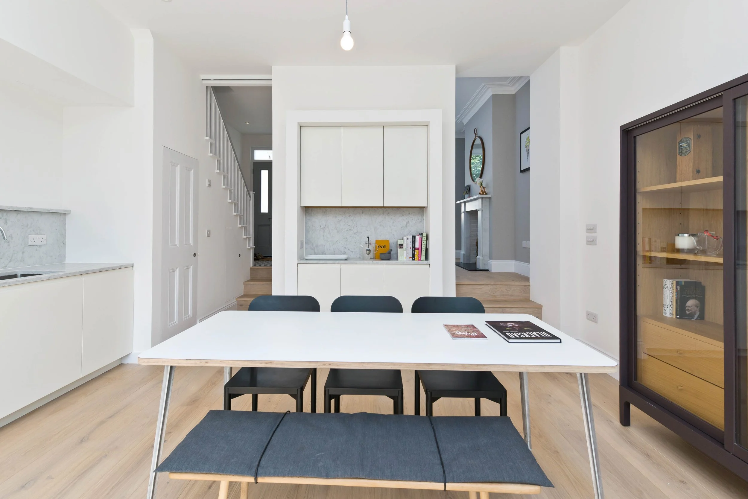 Modern dining room with white table, black chairs, and wooden bench, overlooking a bright kitchen and living area with light hardwood floors.