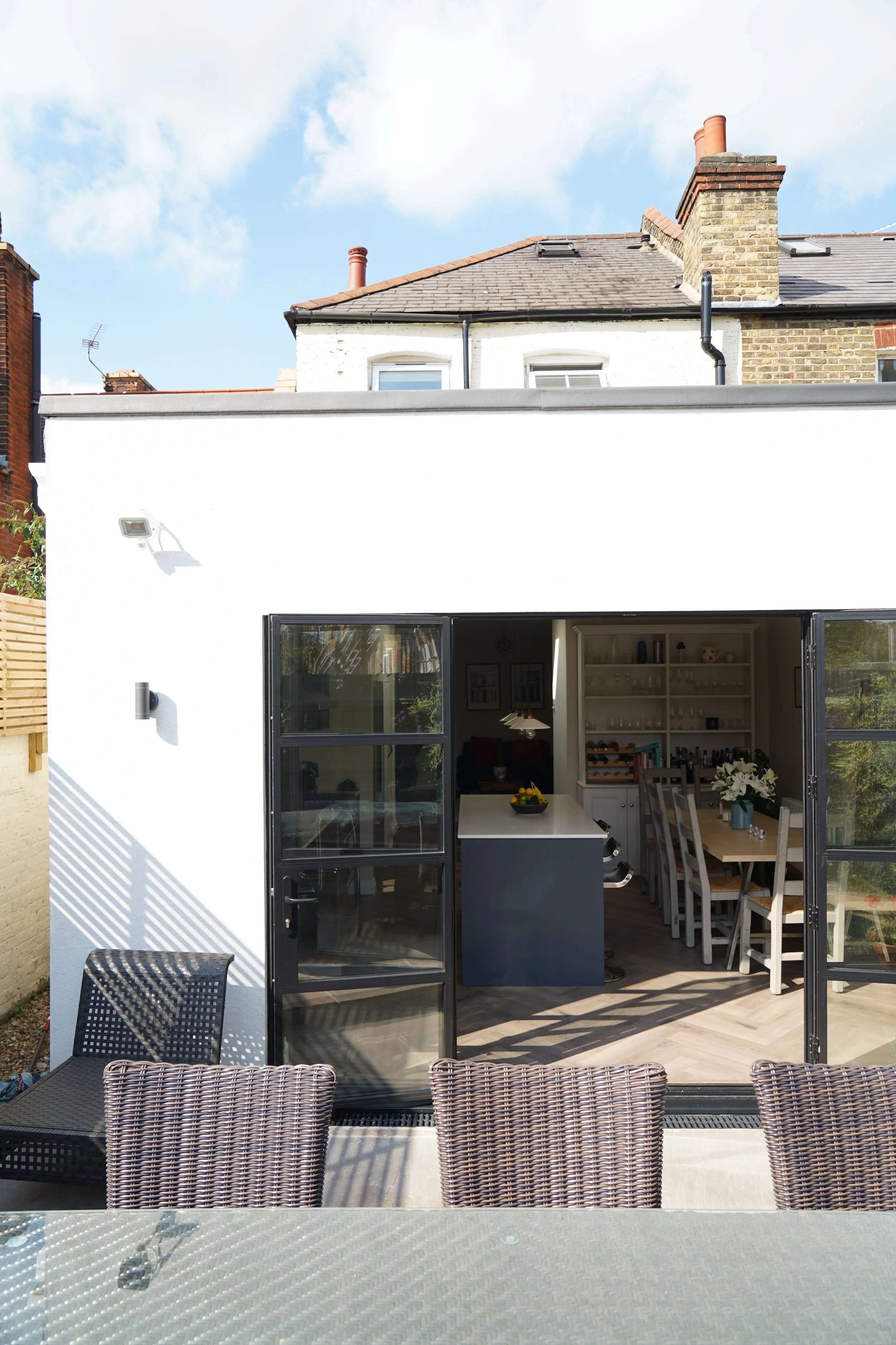 Outdoor patio showing a glass table and chairs in the foreground, with a white building featuring large glass doors opening into a kitchen and dining area in the background, under a cloudy sky.