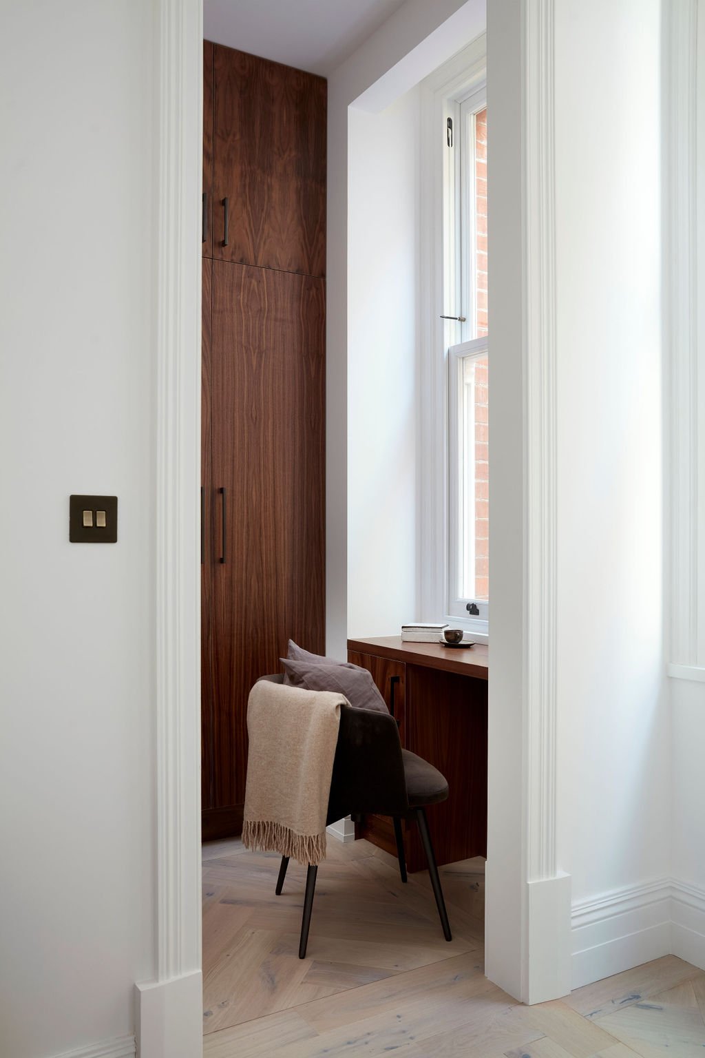 A small built-in wooden desk with a window above it, a dark chair with pillows and a blanket, and a tall wooden cabinet in a bright room.