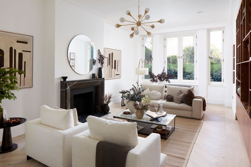Bright living room with white walls, large windows, a white sofa, two white armchairs, a marble coffee table, a black fireplace with a mirror above, abstract wall art, a wooden bookshelf, potted plants, and a modern chandelier.