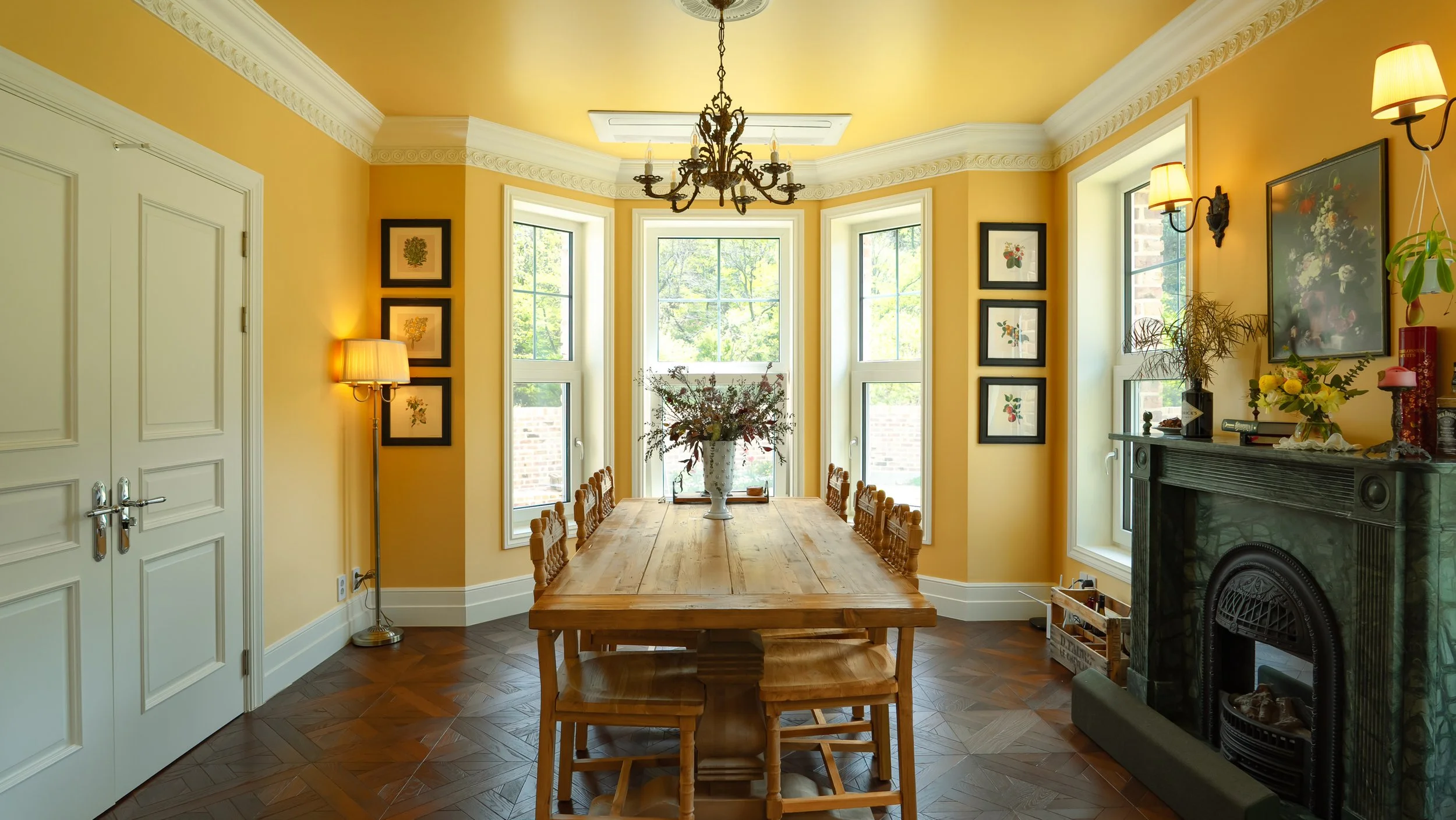 Sunny dining room with a large wooden table, nine chairs, a chandelier, and a floral arrangement in a white vase, surrounded by yellow walls with decorative crown molding, framed pictures, and windows letting in natural light.