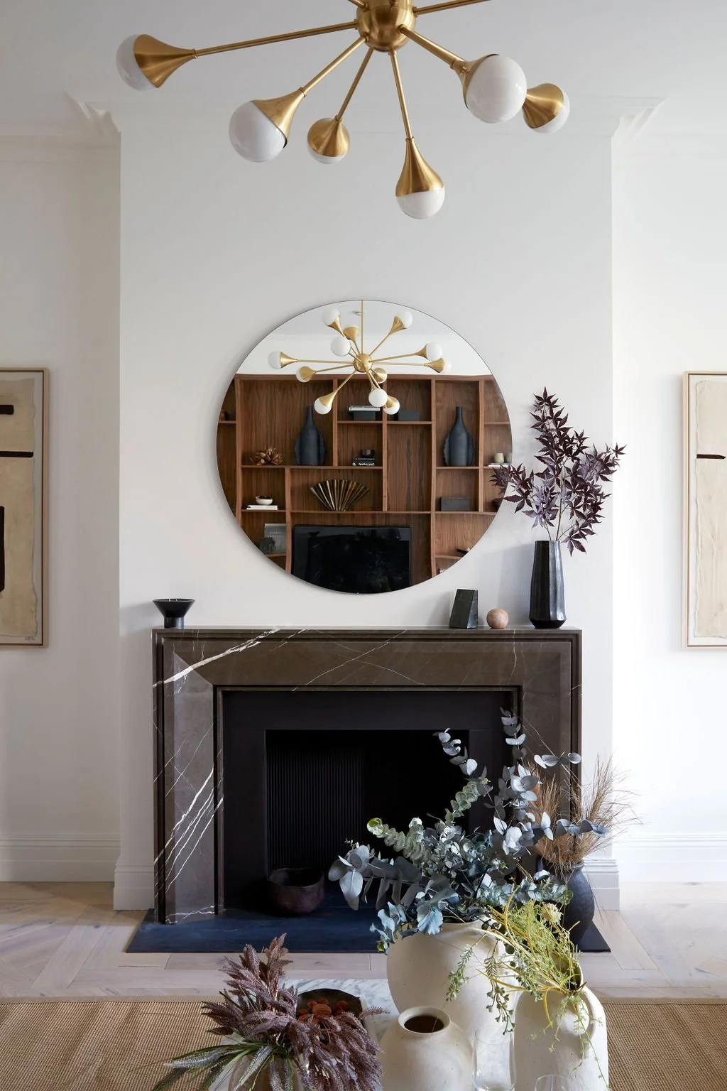 Living room with a decorative black fireplace, a round mirror above it reflecting a wooden bookshelf, a modern gold chandelier, and a vase with dark purple leaves on a side table. Potted plants in the foreground.