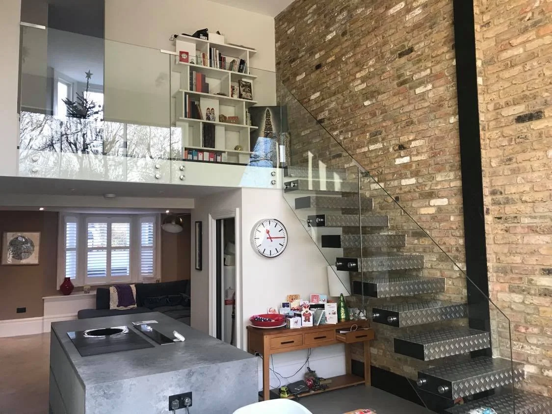Interior view of a modern house with an open loft, exposed brick wall, a staircase with metal steps and glass railing, a large wall clock, and a small table with various items on it.