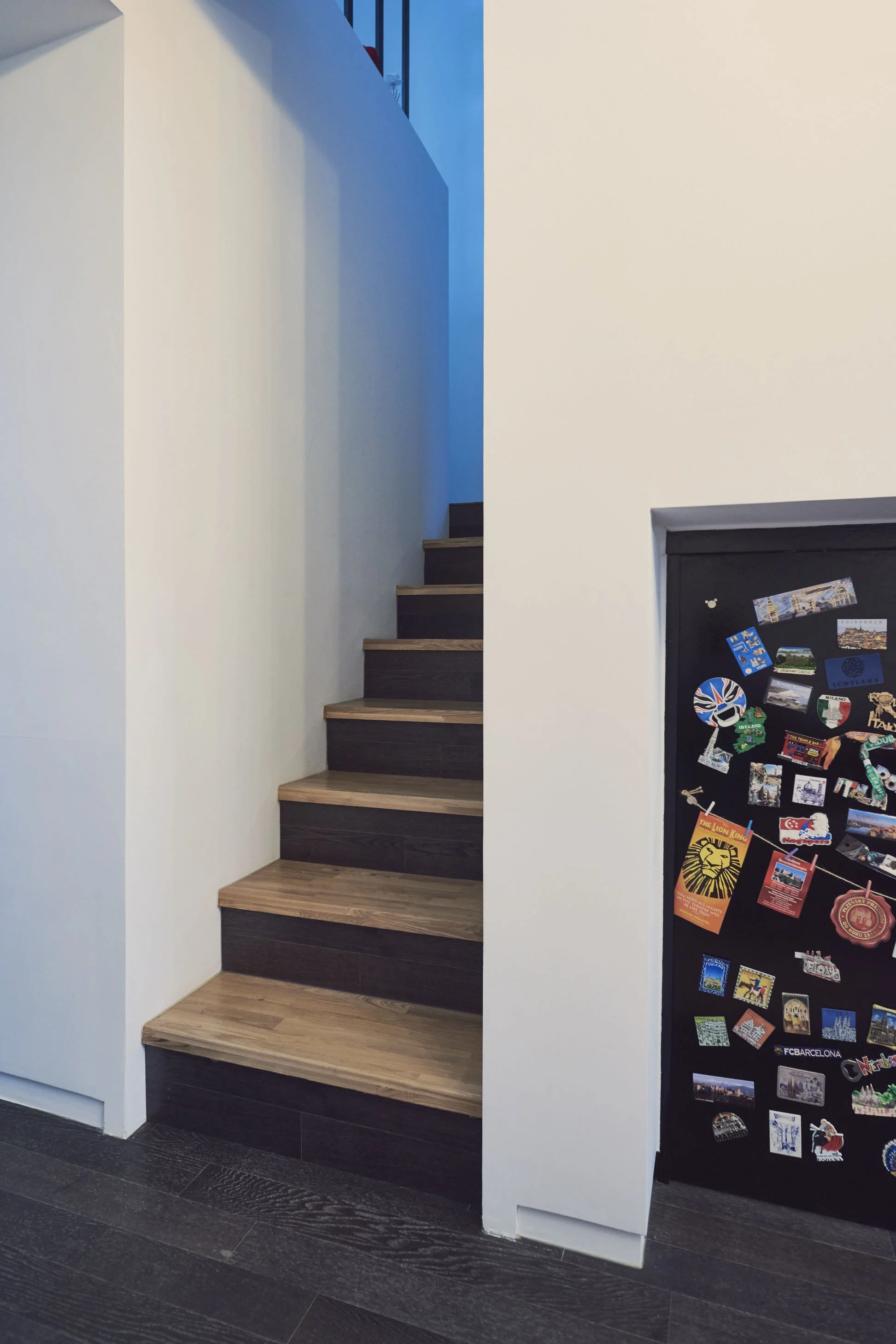 Indoor wooden staircase with dark steps and light wooden treads next to a white wall, adjacent to a black refrigerator covered with colorful magnets and stickers.