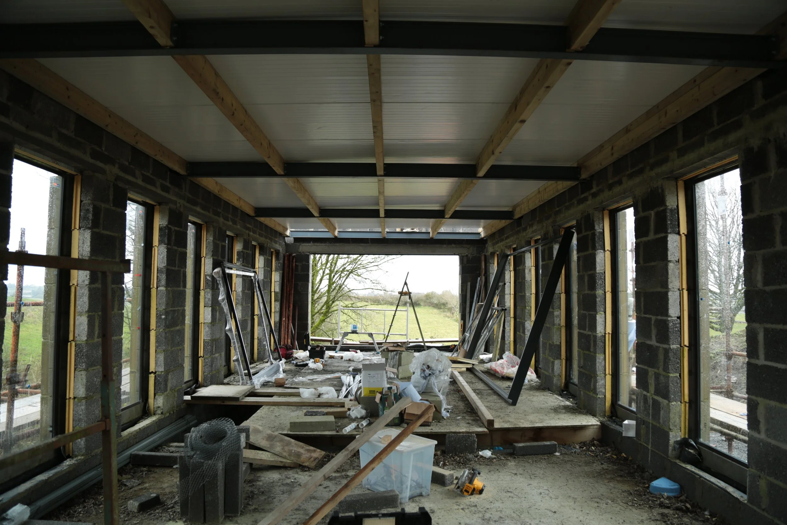Interior of a building under construction with unfinished brick walls, large window openings, and construction tools and materials scattered on the floor.