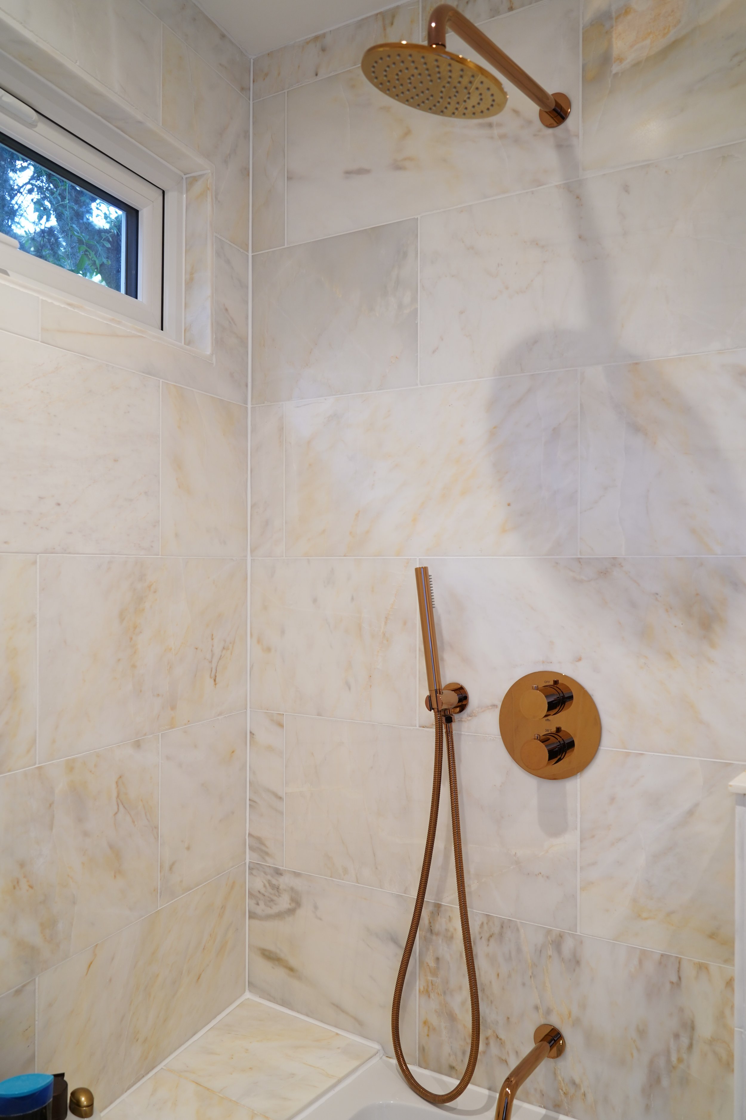 Marble-tiled shower with a gold showerhead, a handheld shower wand, and a control panel, with a small window at the top showing trees outside.