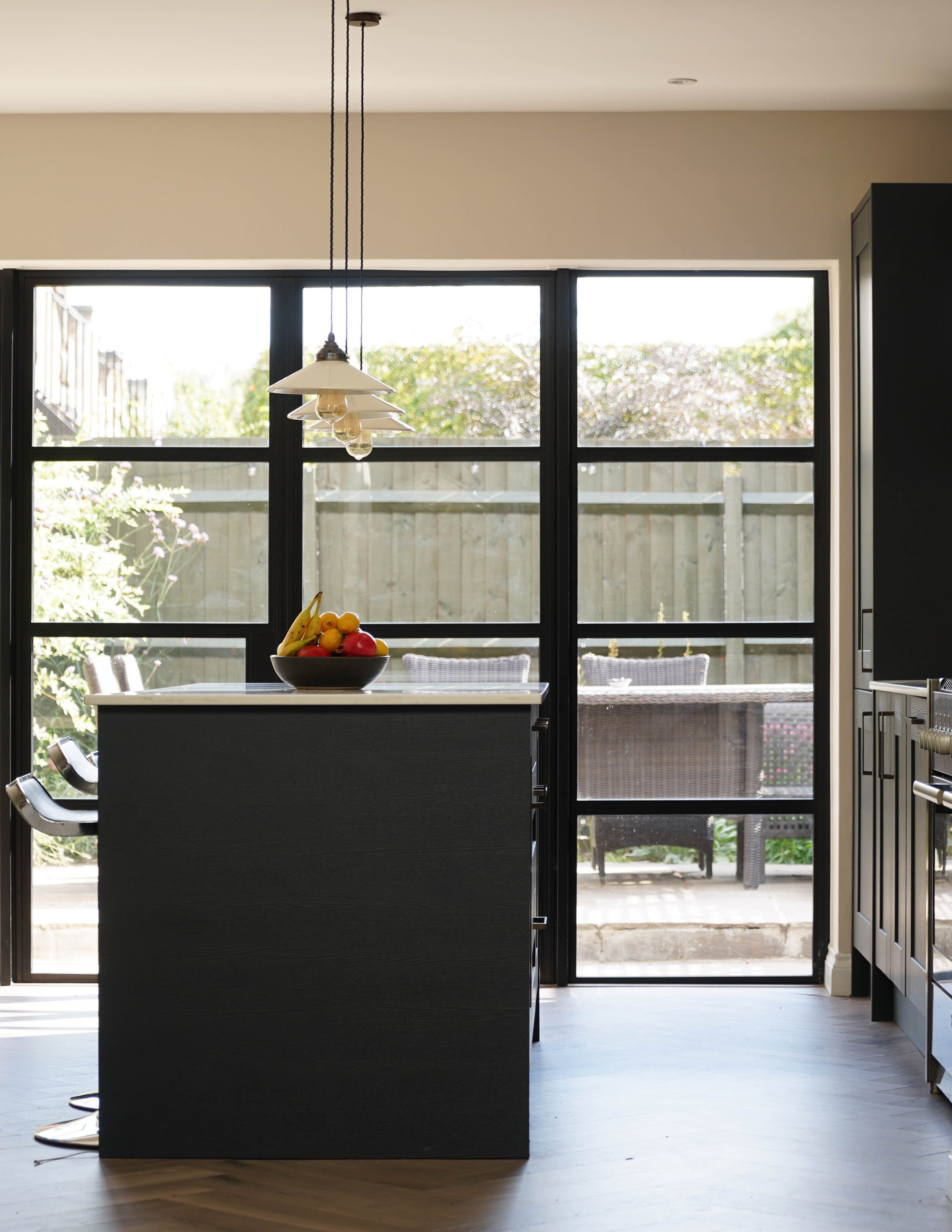 Modern kitchen with black cabinetry, a black kitchen island with a fruit bowl, and a large window with black framing opening to an outdoor patio with chairs and greenery.
