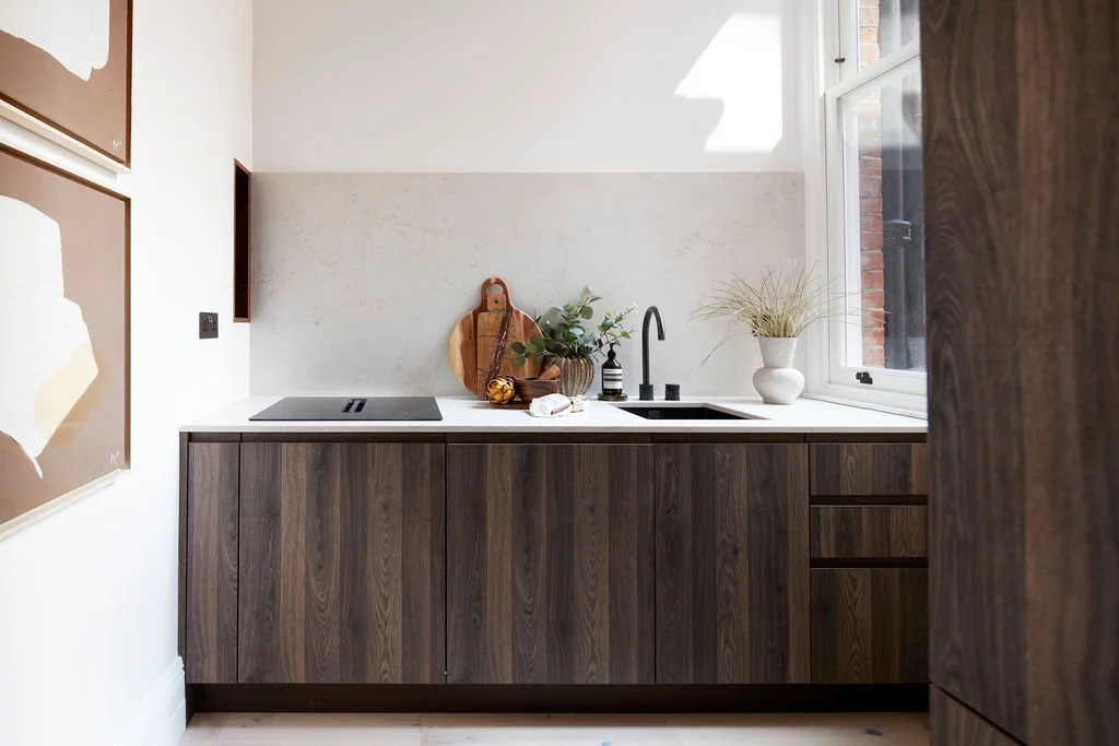 Modern kitchen with dark wood cabinets, white countertop, black faucet, sink, potted plant, and window letting in natural light.