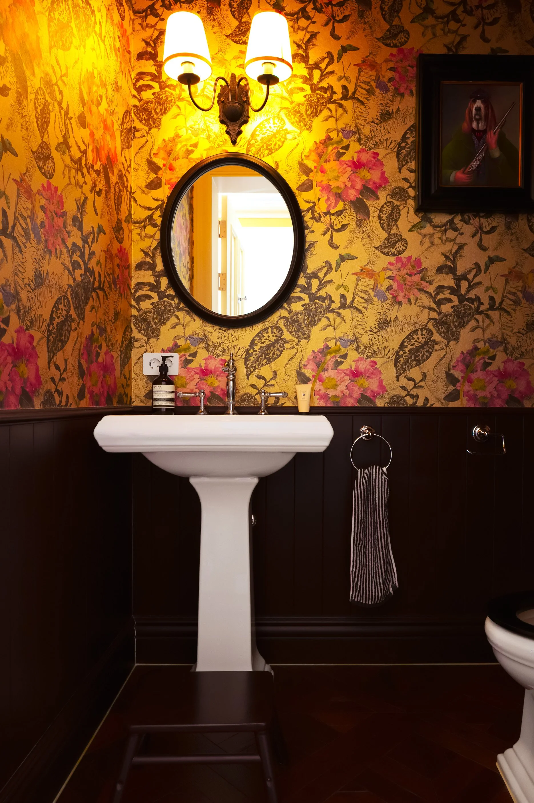 A bathroom with wallpaper featuring pink and yellow flowers and dark leafy patterns, a white pedestal sink with silver faucets, a round black-framed mirror, two wall-mounted light fixtures with white shades, a framed cartoon dog painting, and a strip