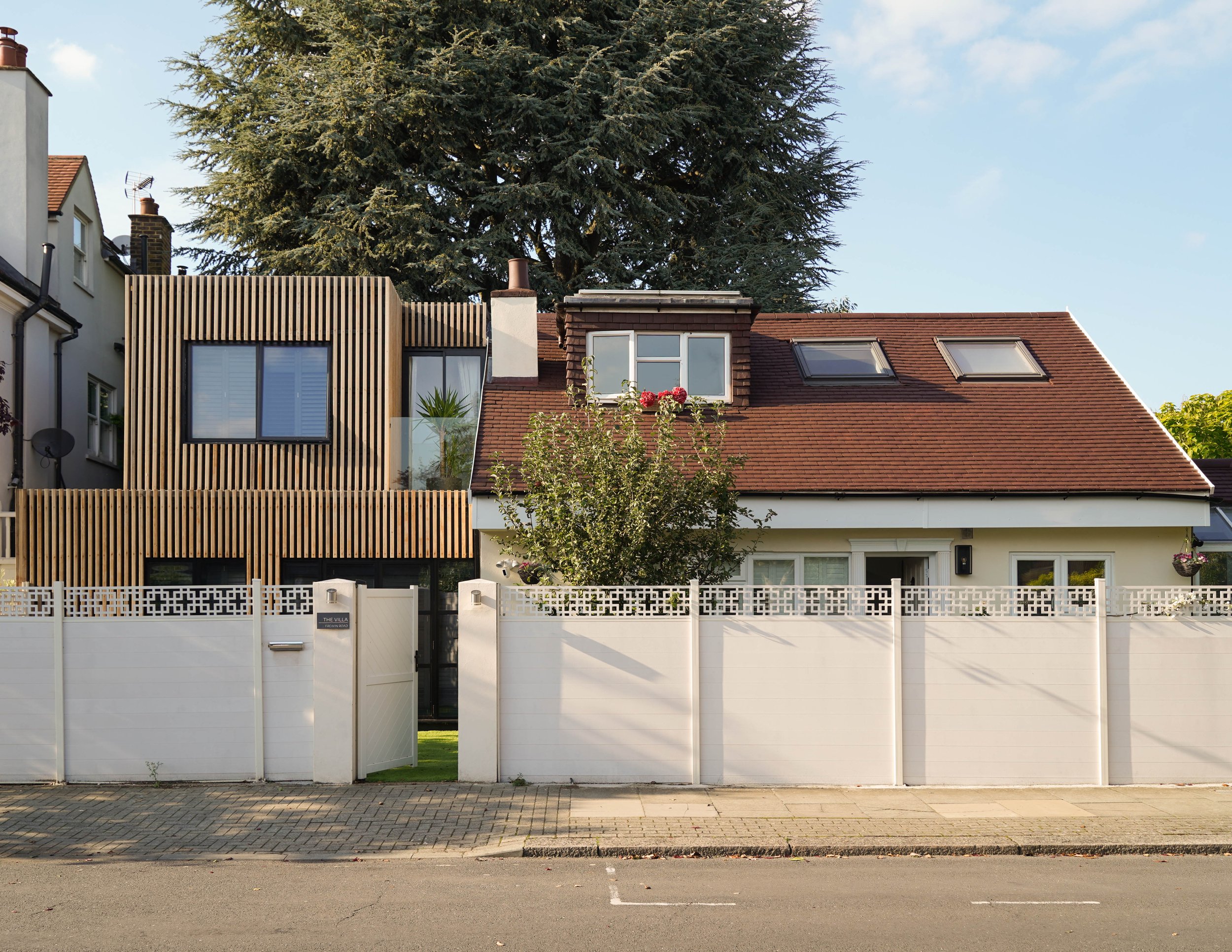 A residential house with a modern wooden extension on the left and a traditional roof with skylights on the right, surrounded by a white fence and a tree in front.
