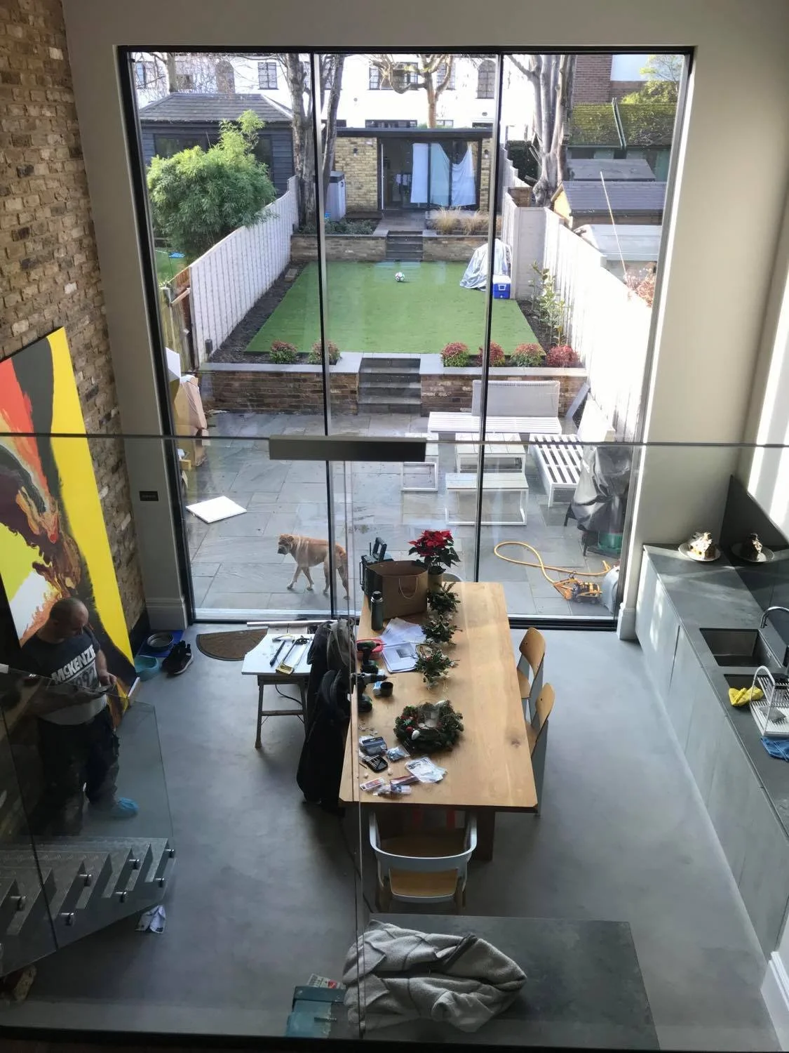 View from inside a modern kitchen looking out through a large glass sliding door to a backyard with a small lawn, patio furniture, and a dog.