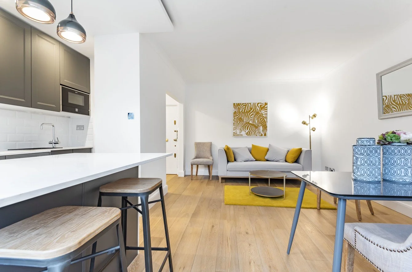 Modern living room with a white sofa, yellow and gray pillows, a abstract gold and white painting, a white wall, wooden floor, a gray table with blue patterned boxes, a dining table, and a kitchen area with gray cabinets and a white countertop.