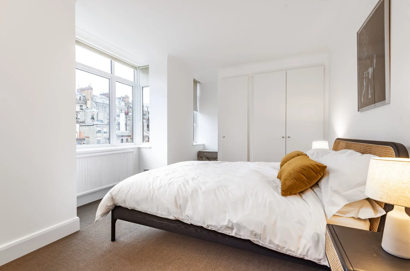 Bright minimalist bedroom with large windows, white walls, a bed with white linens and two mustard-colored pillows, and a nightstand with a lamp.