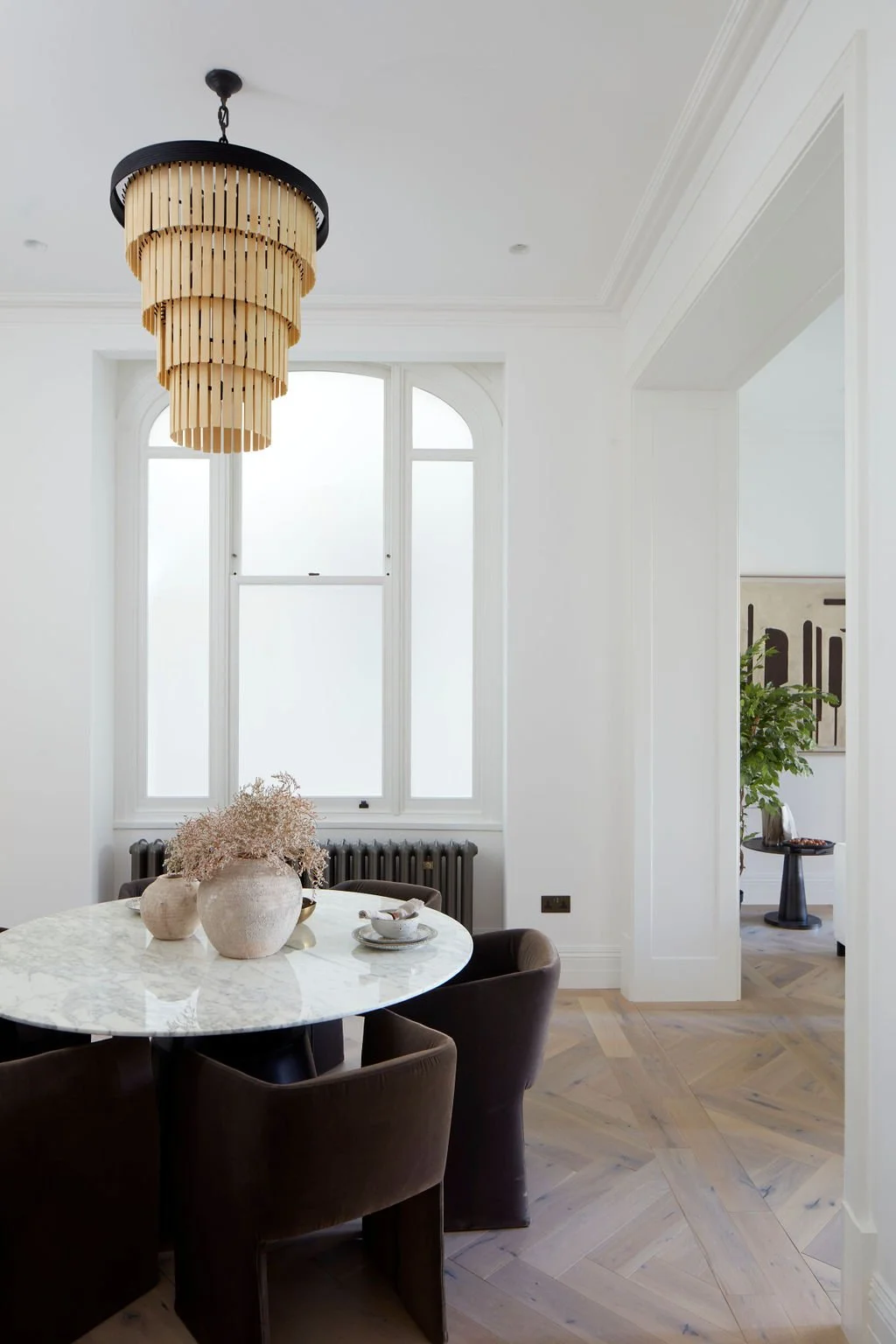 Dining room with a round marble table, dark upholstered chairs, and a chandelier with wooden slats hanging from the ceiling. Large windows allow natural light into the space.