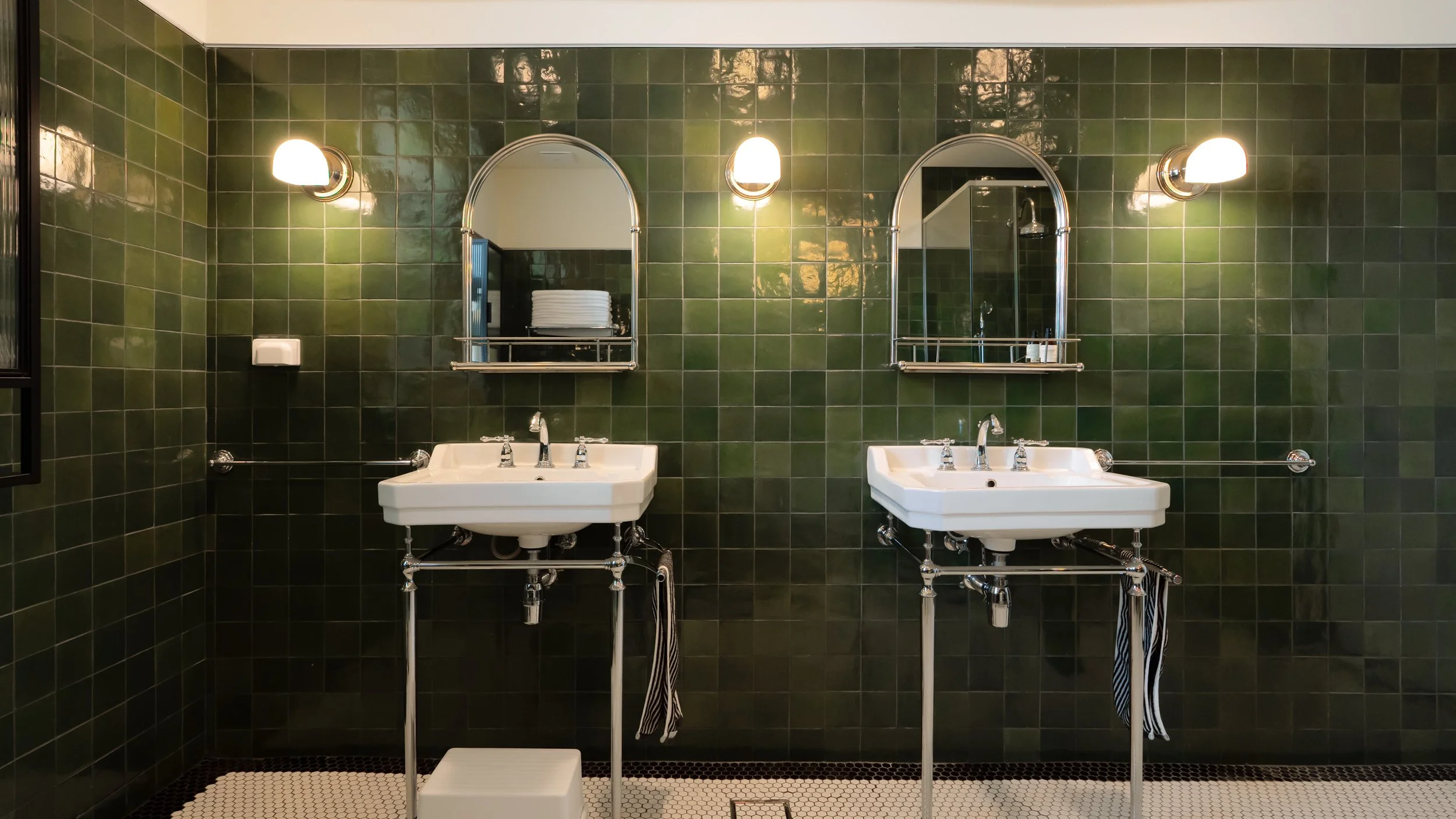 Bathroom with two white sinks and twin arched mirrors on a dark green tiled wall.