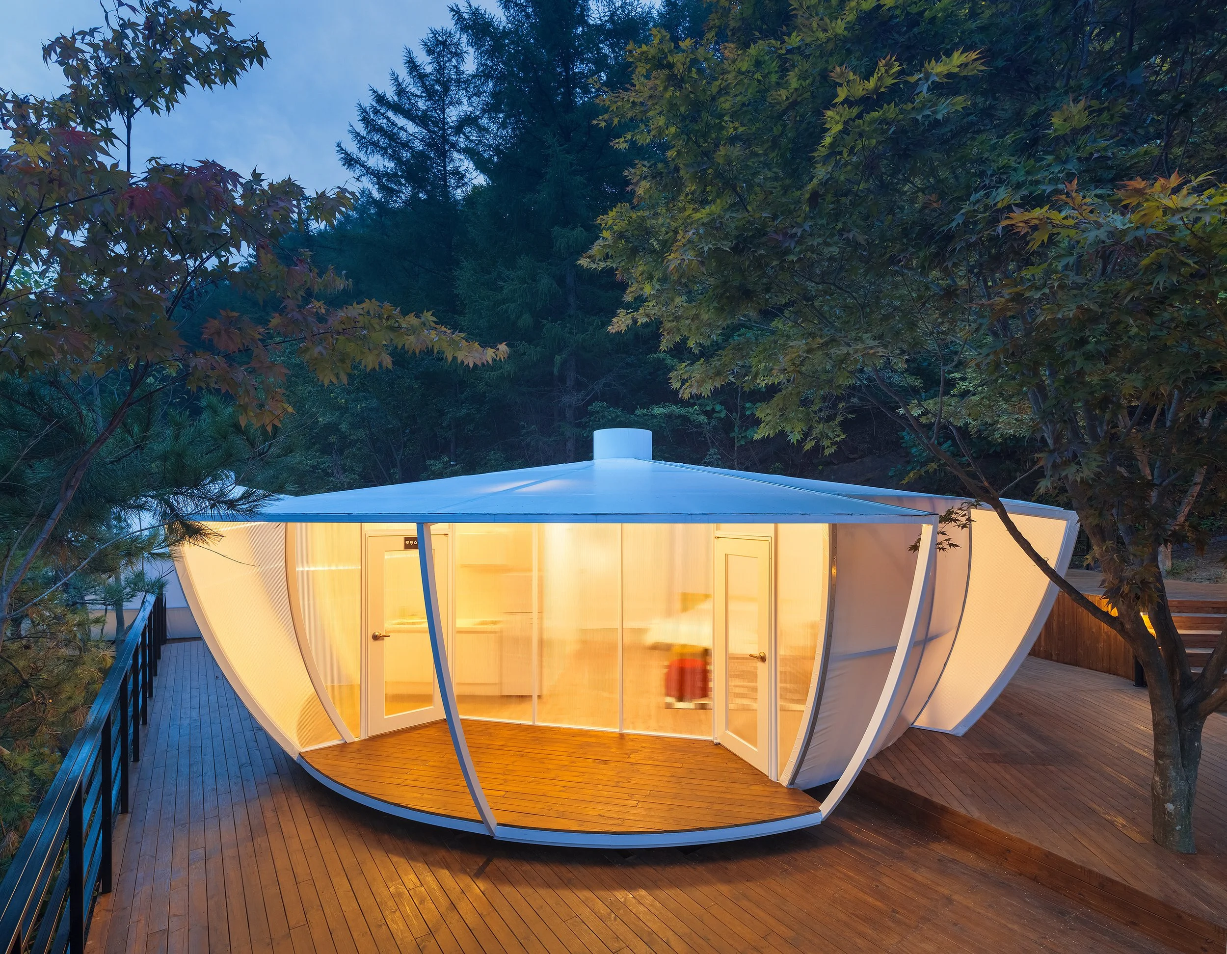 A modern, circular glass house with illuminated interior, situated on a wooden deck surrounded by trees, during dusk.