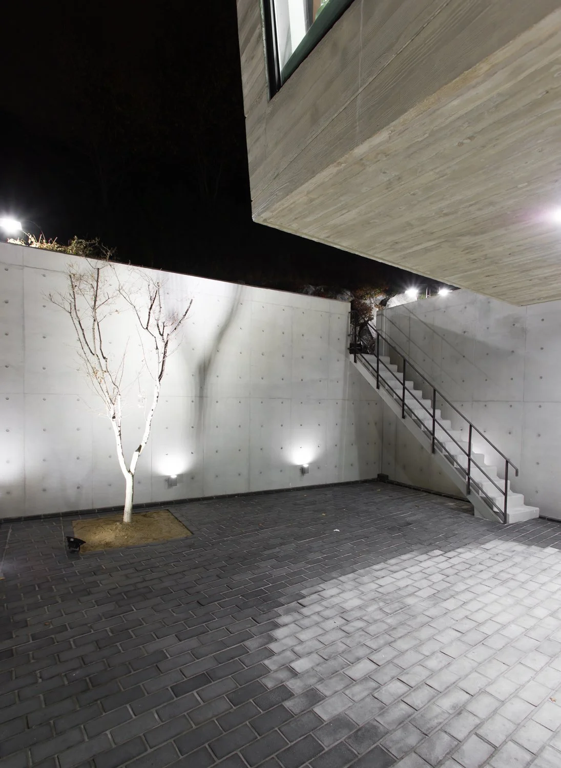 Nighttime scene of a minimalist outdoor space with a single leafless tree, concrete walls with installed lighting, brick pavers on the ground, and a metal staircase leading to an upper level, all illuminated by outdoor lighting.