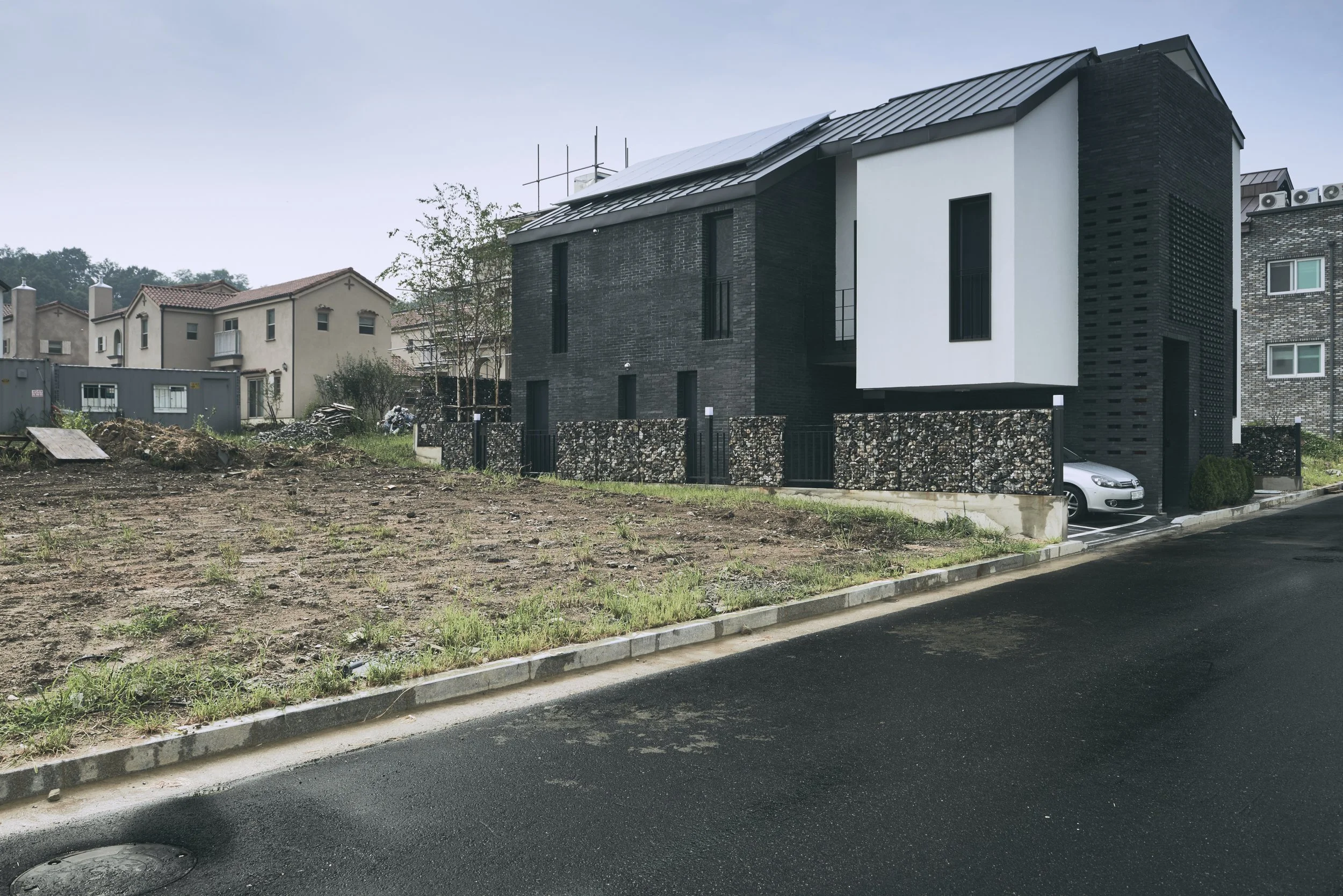 Modern residential house with black and white exterior, situated next to a street, with a driveway under a carport and a small yard with a brown patch of dirt and some grass.