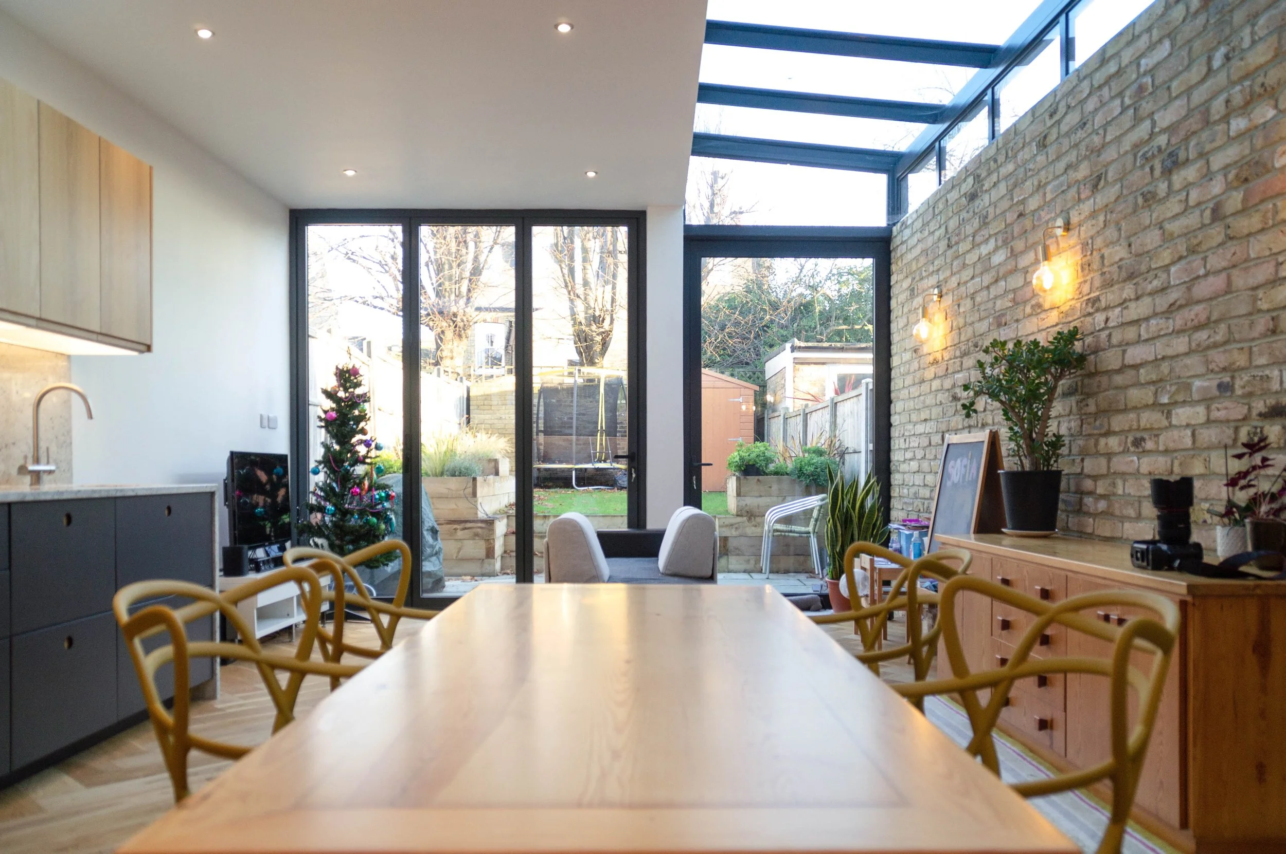 Modern dining room and kitchen area with glass doors leading to a backyard, decorated for Christmas with a small tree, sitting area with chairs, brick wall with lighting, and plants.