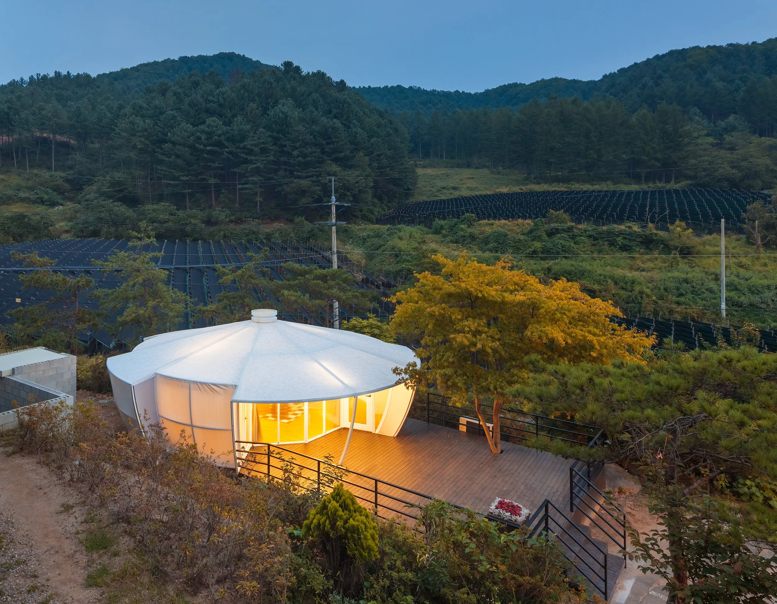 A modern, illuminated building with a large, umbrella-like roof on a wooden deck, surrounded by trees and hillside farmland with dark crops, during dusk.