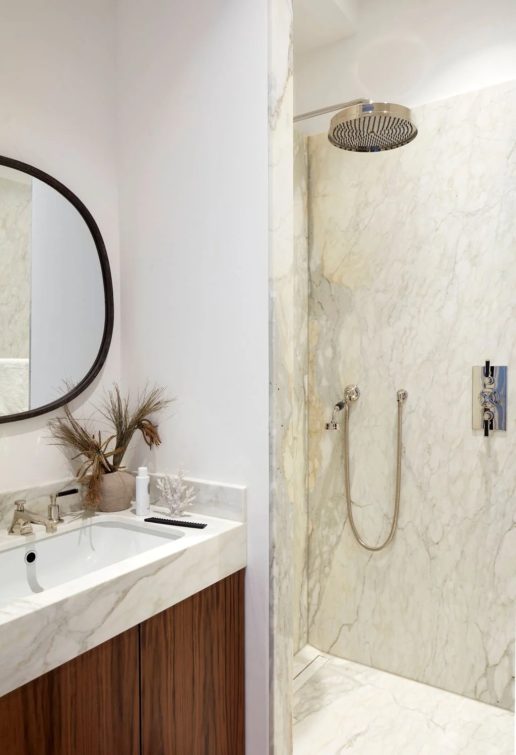A bathroom with a marble shower area and a vanity with a mirror, decorative dried plants, and a coral ornament.