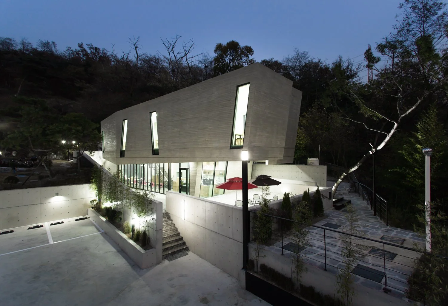 Modern multi-story building with large angular windows, concrete exterior, and outdoor patio with red and black umbrellas, surrounded by trees at dusk.