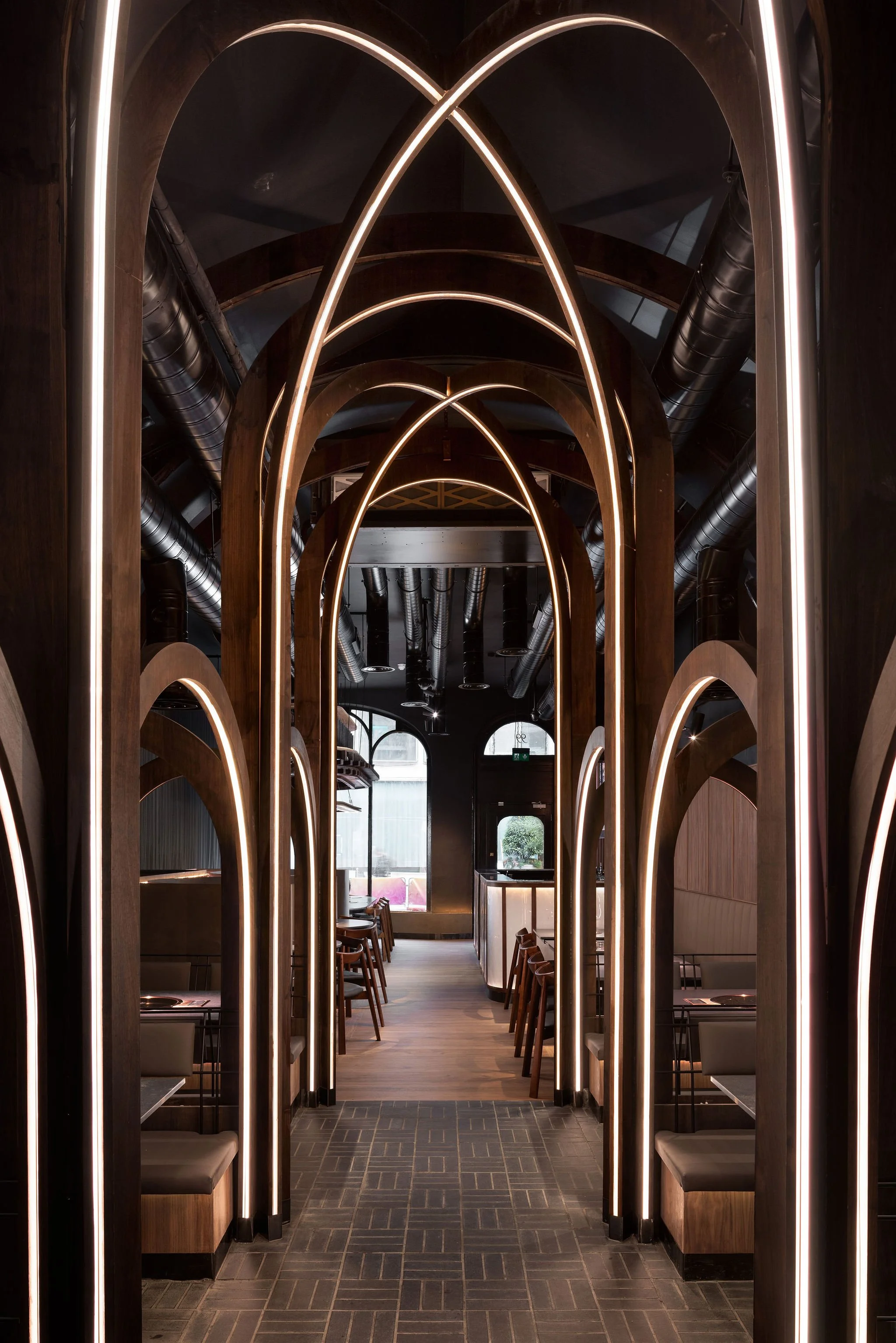Modern restaurant interior with wooden archways and integrated lighting, featuring dining booths on either side and a hallway leading to a seating area near windows.