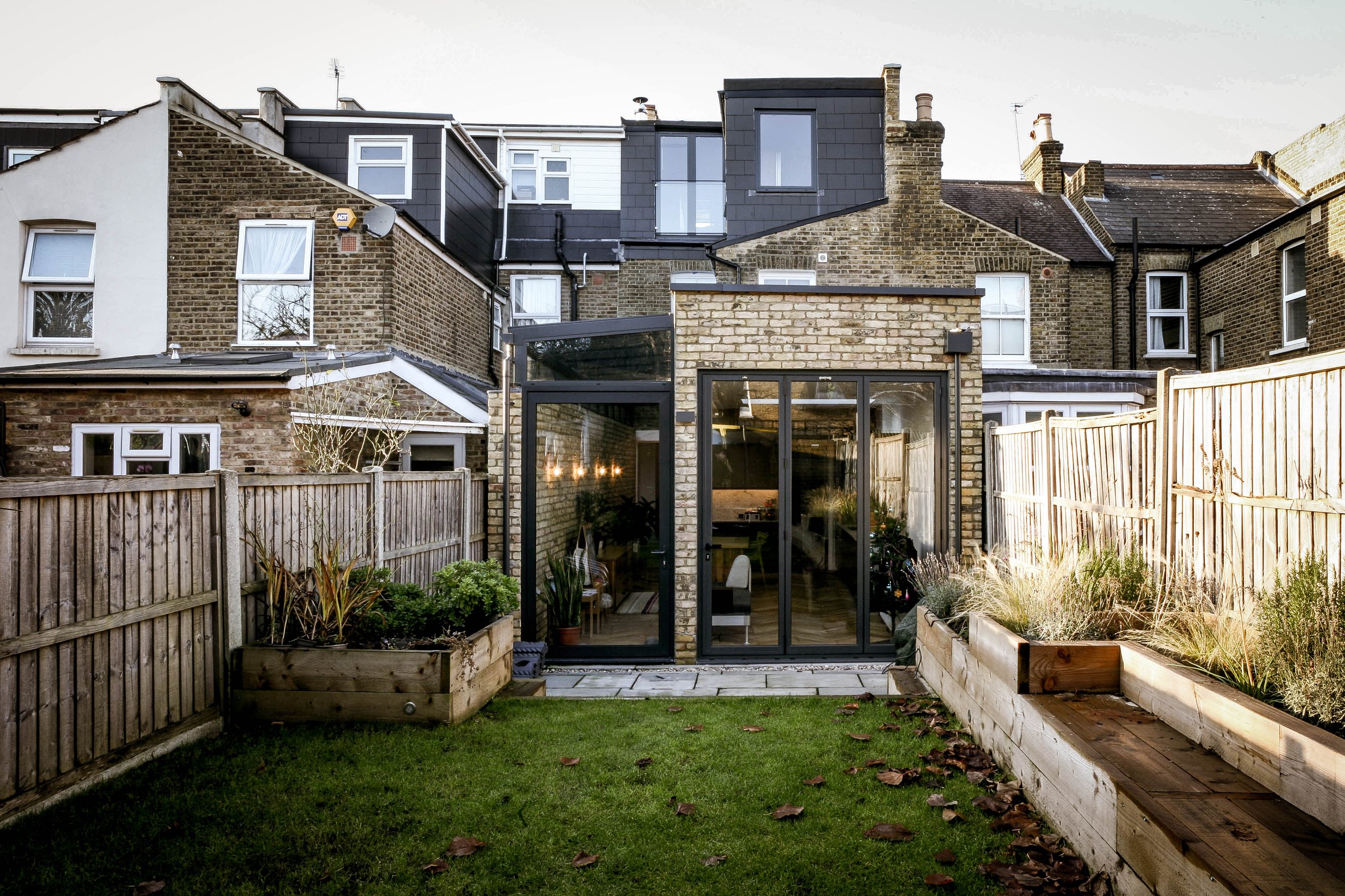 Backyard garden with green grass, wooden fence, and modern glass extension on brick house showing interior dining area with chairs and hanging lights.