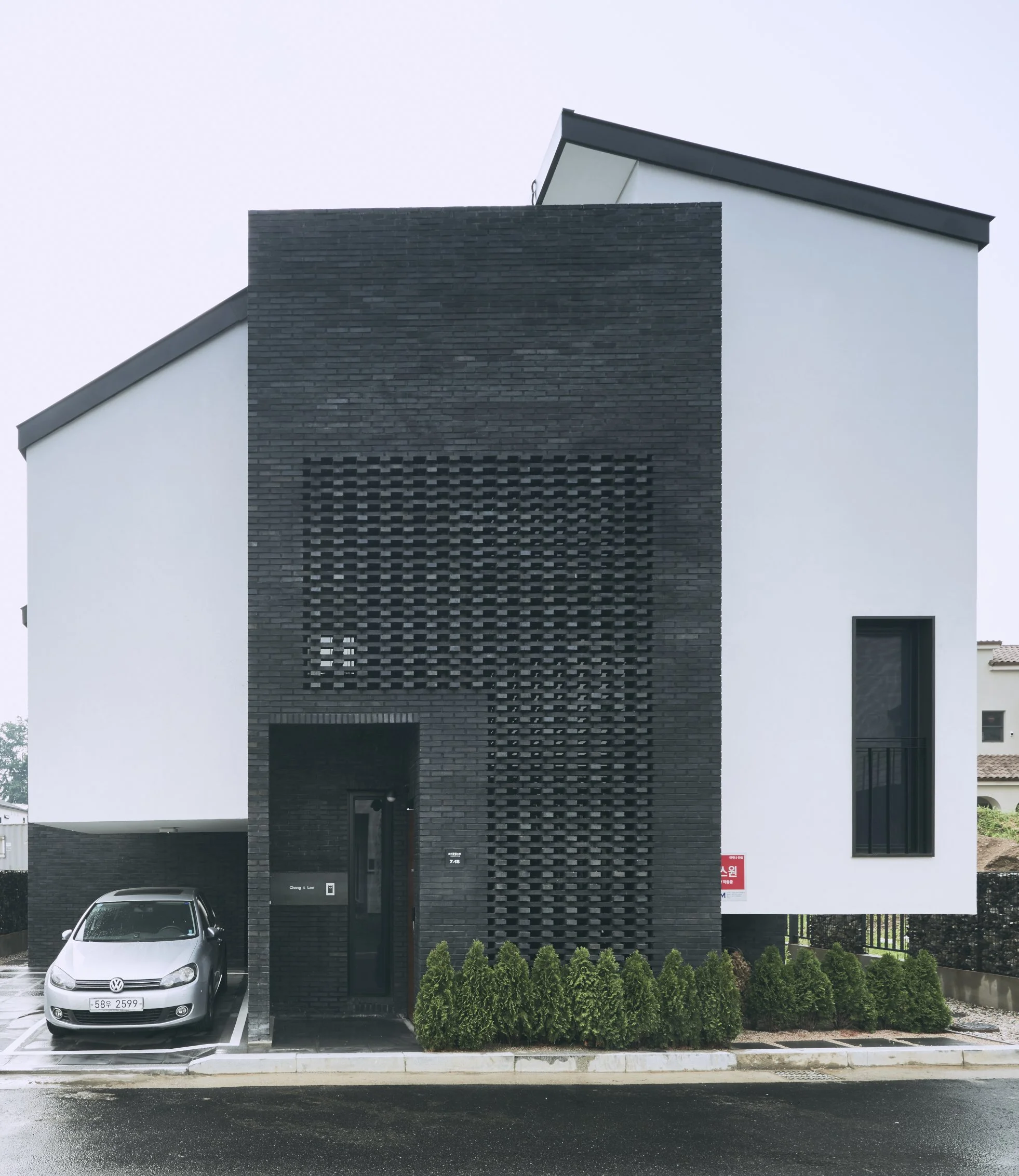 Modern house with black brick facade, white exterior walls, a small parking spot with a silver car, and neatly trimmed bushes in front.
