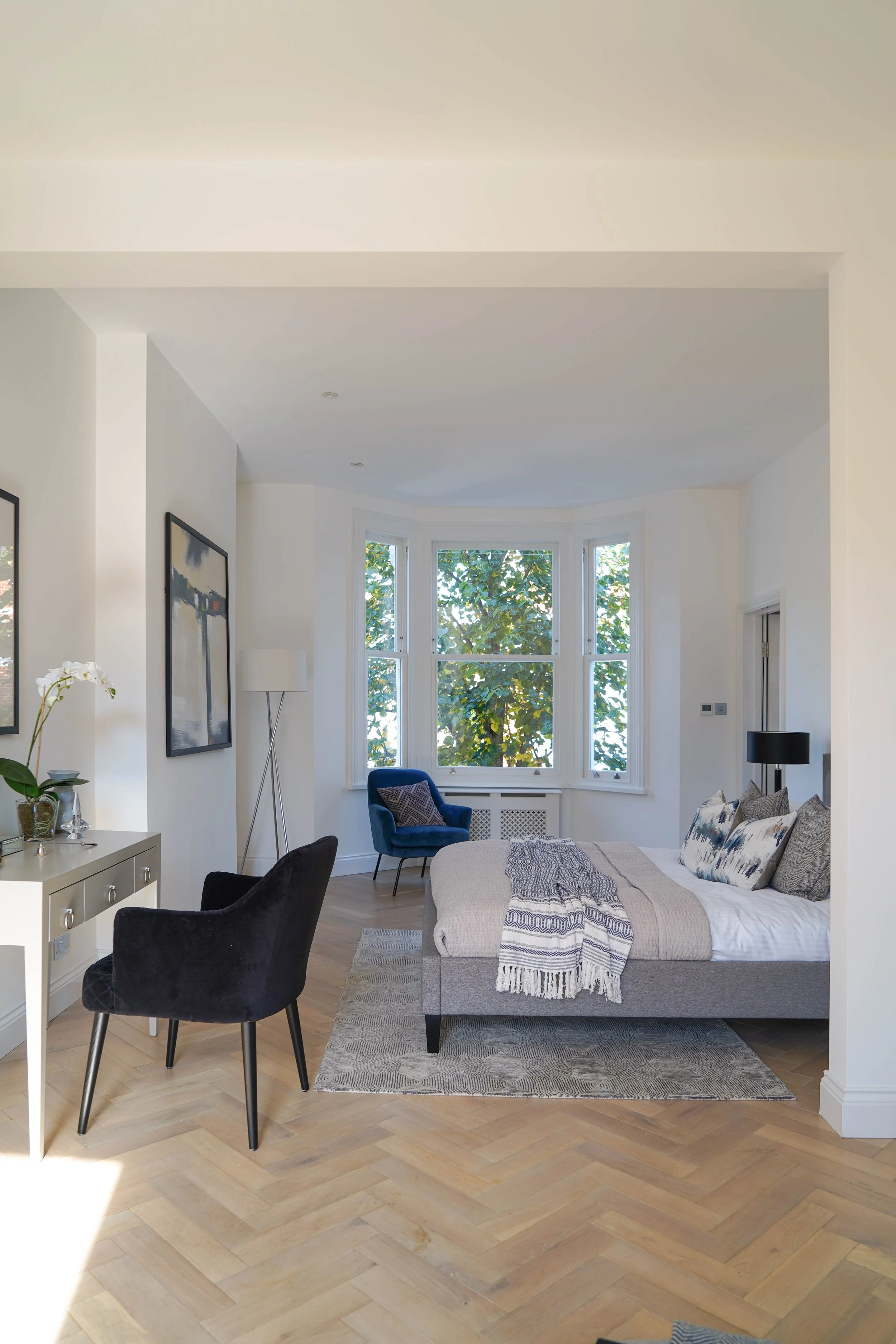 Bright bedroom with large bay window, gray bed with patterned pillows and throw, blue armchair, black side table with lamp, black chair, white dresser with orchid, abstract paintings, hardwood floor, and modern decor