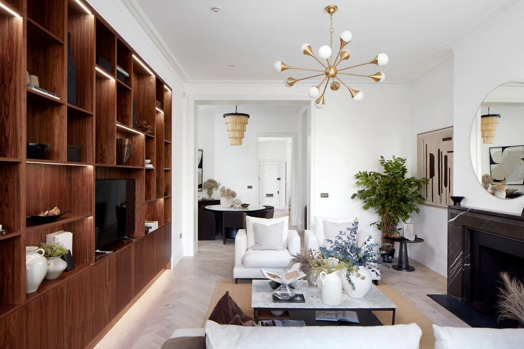 Living room with white couches, a black fireplace, and a wooden bookshelf with decorative objects and books. There is a coffee table with vases and plants. A large green plant stands next to a round mirror on the wall. A modern chandelier hangs from 