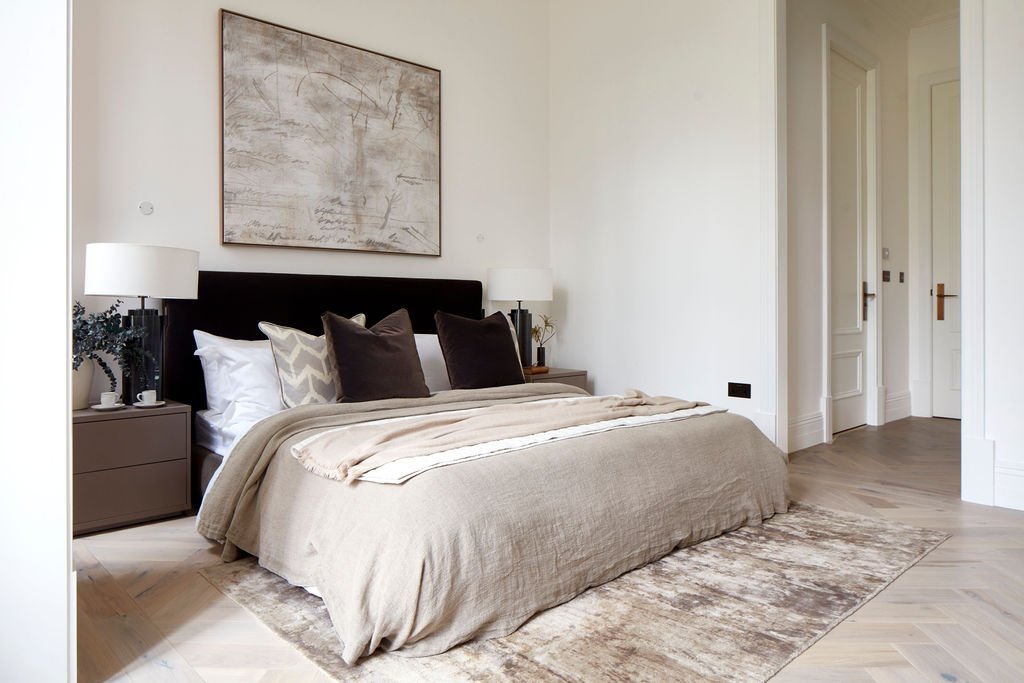 Modern bedroom with beige bedspread, four throw pillows, two side tables with lamps, wall art, and wooden floors.