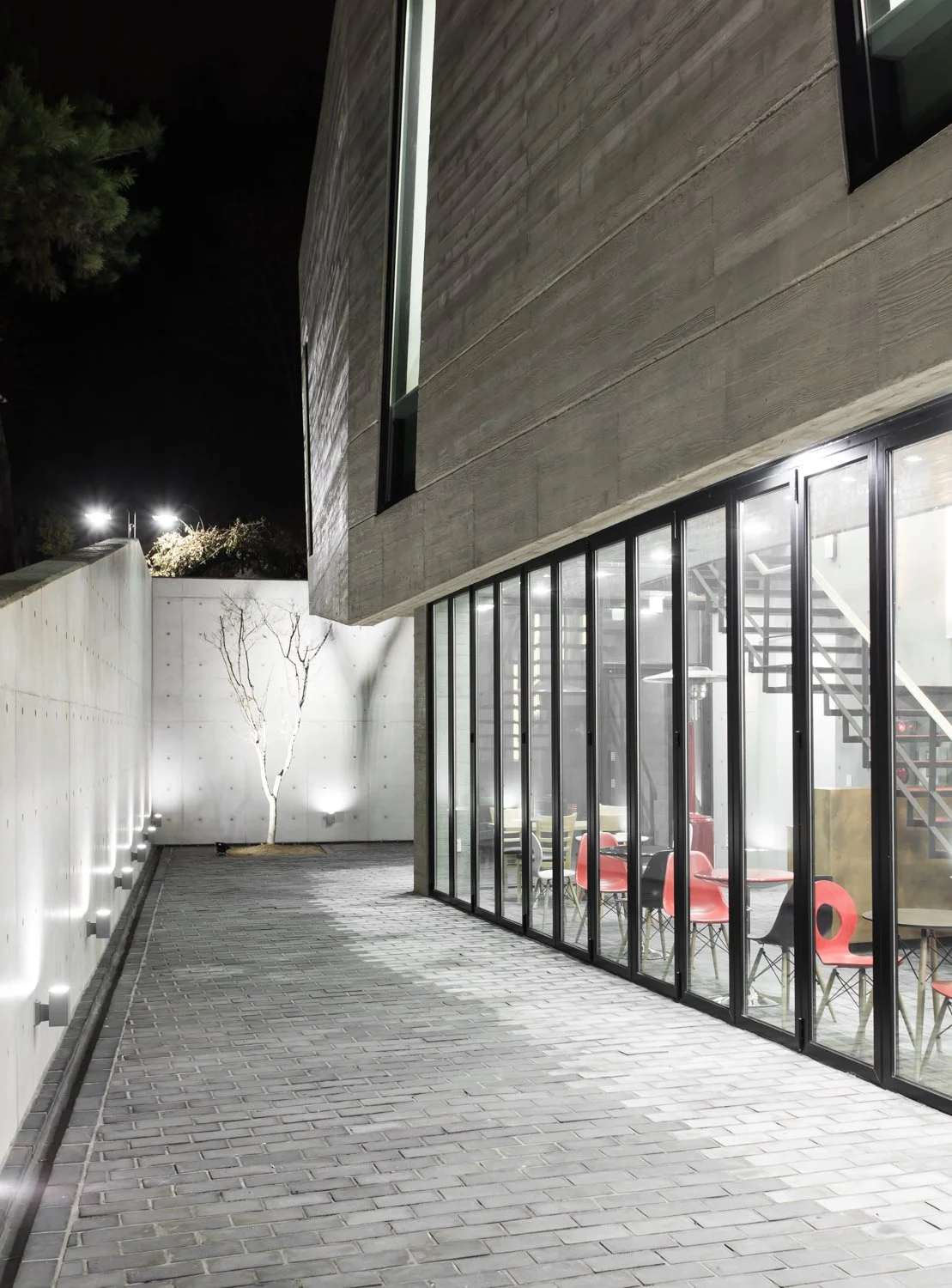 Night view of a modern building with large glass windows, a brick walkway, a white concrete wall with small lights, and a small leafless tree in the patio area.
