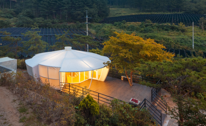 Unique modern house with a curved design, large windows, and a roof with a central vent, situated on a hillside with surrounding trees and a viewing deck at dusk.