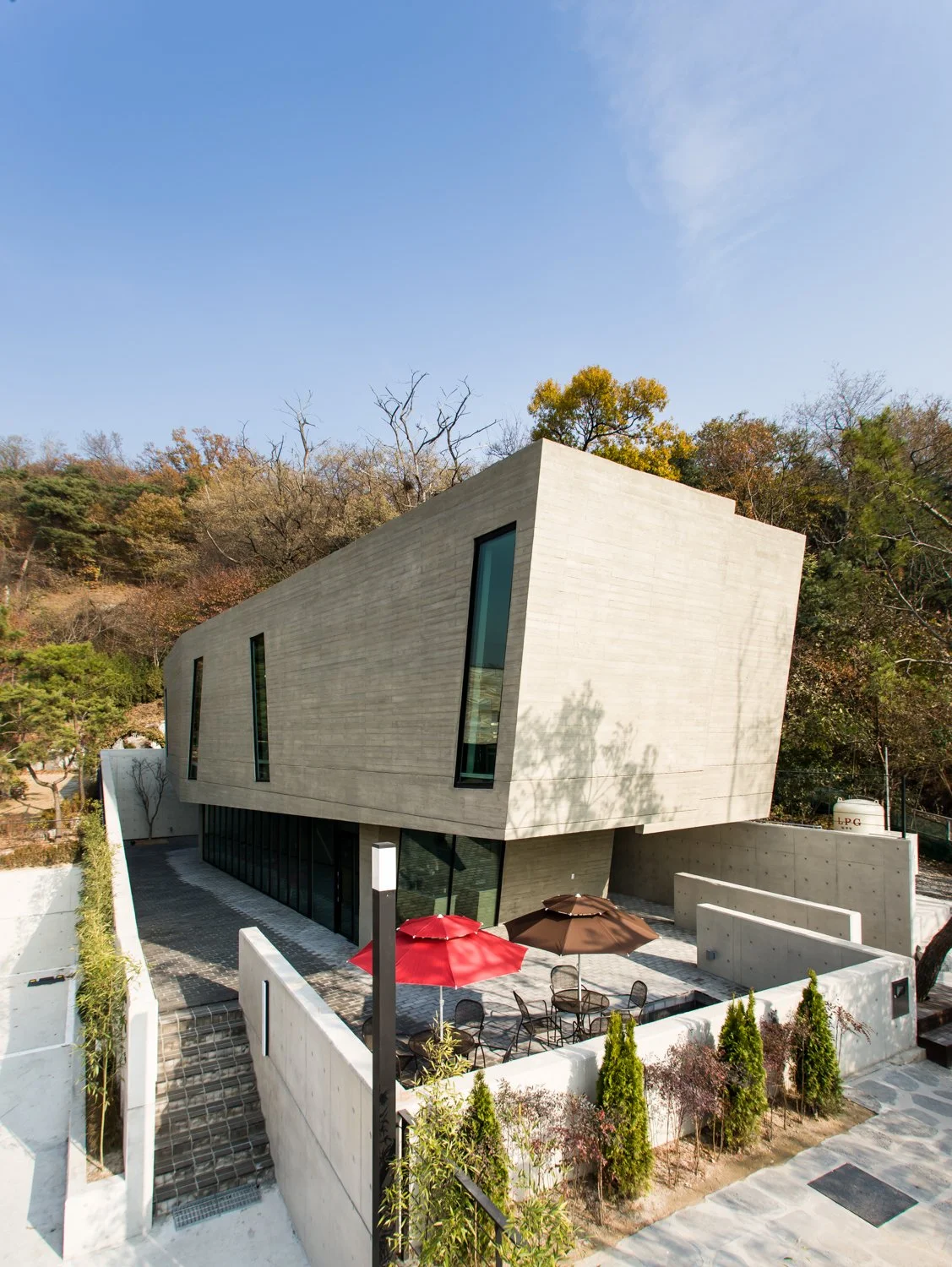 Modern concrete building with angular design and large windows, outdoor patio with umbrellas, surrounded by trees and landscaping, under clear blue sky.
