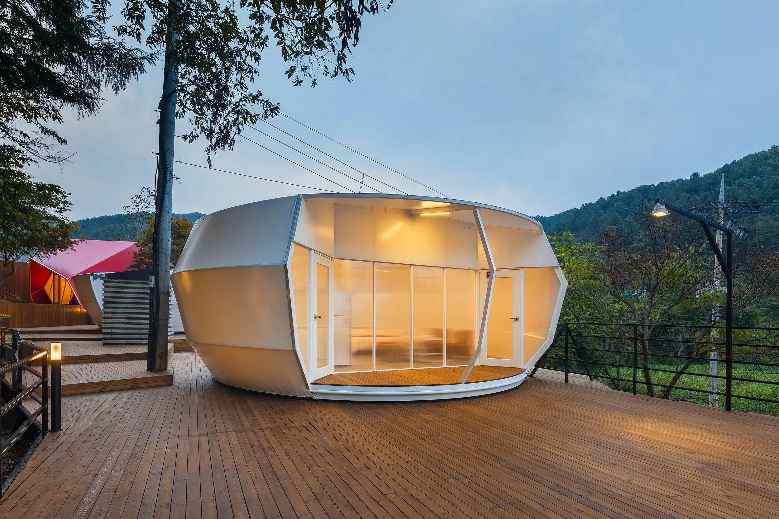 Modern, oval-shaped building with large glass doors on a wooden deck, illuminated from within, surrounded by trees and mountains at dusk.
