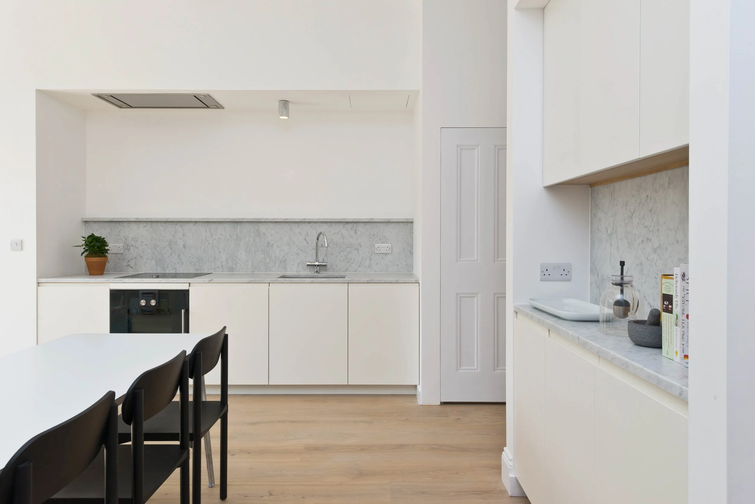 Modern minimalist kitchen with white cabinets, marble countertops, a small potted plant, black dining chairs, and wooden flooring.