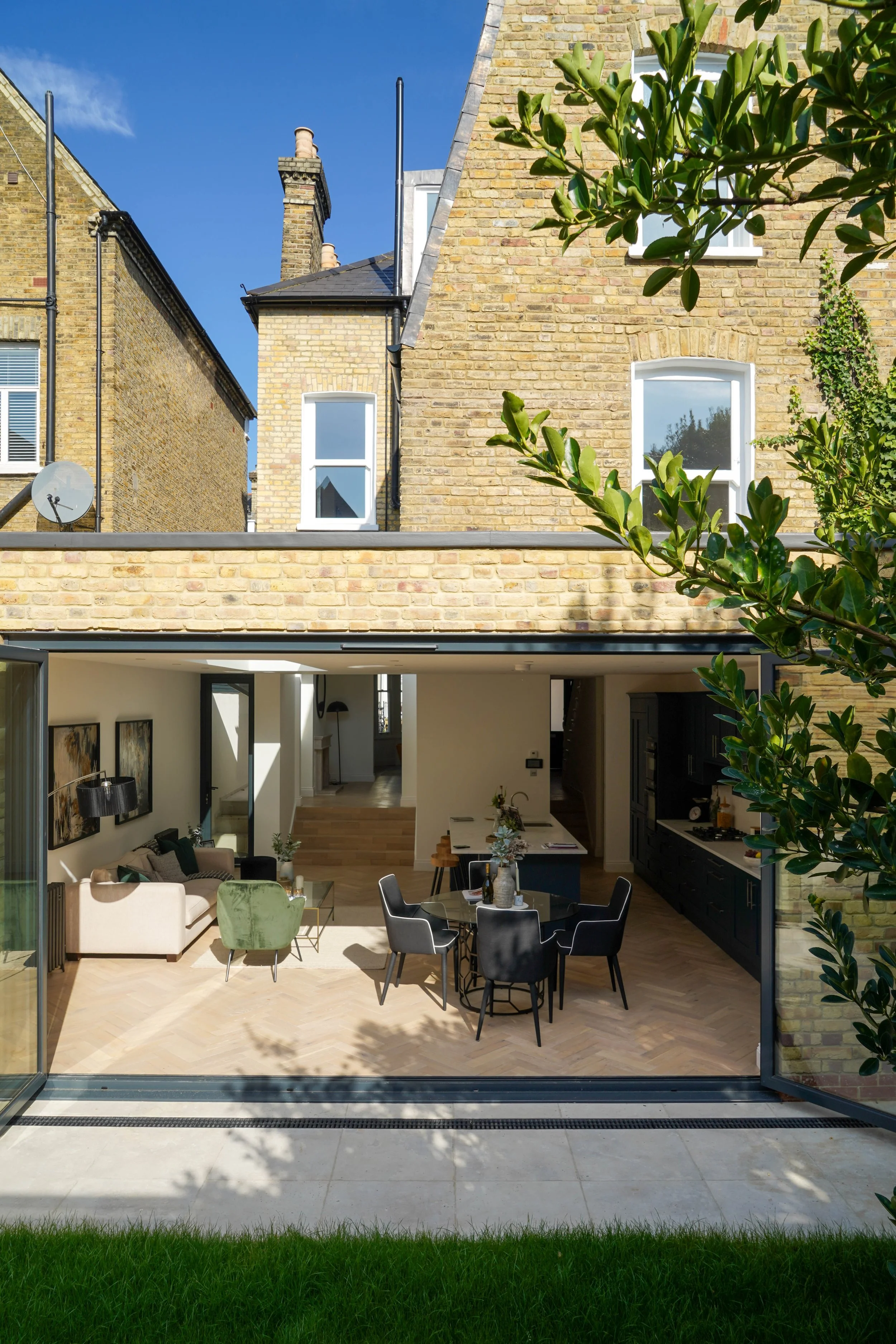 Interior view of a modern open-concept living and dining area with large glass sliding doors opening to a green backyard, featuring a round glass dining table with black chairs, a beige sofa, a green armchair, and black kitchen cabinets.