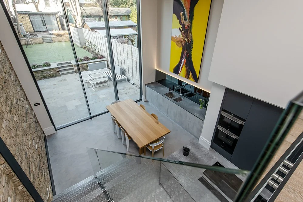 From an elevated view, a modern kitchen with a wooden dining table and chairs, a large abstract painting, floor-to-ceiling glass doors leading to an outdoor patio, and a brick wall on the left side.