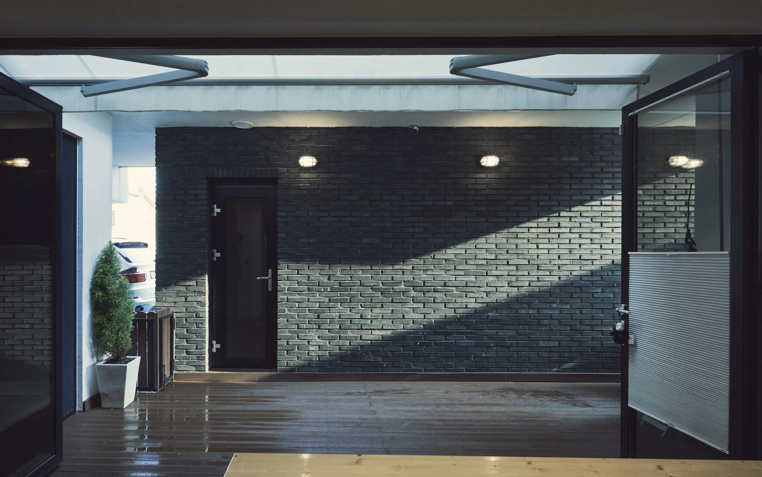 Indoor patio with wooden flooring, black brick wall, glass door, potted plant, and exterior view of a car.
