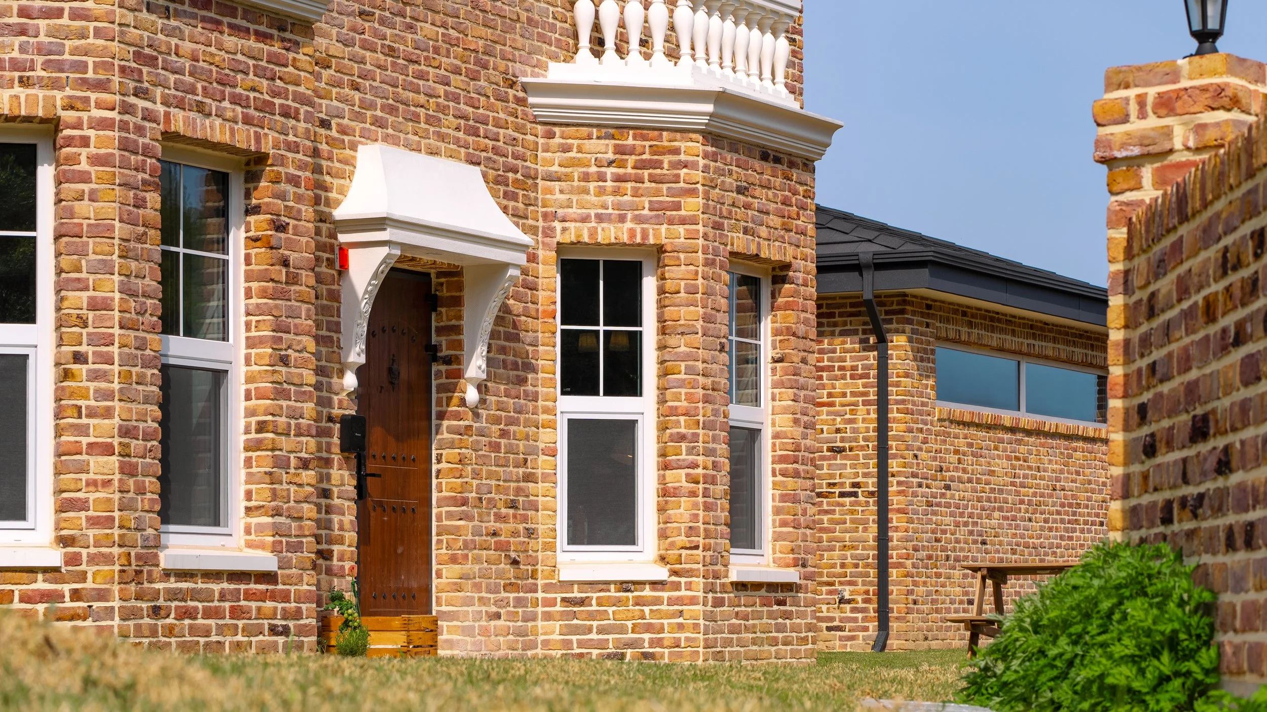 A red brick building with white-framed windows, a wooden front door with decorative black hardware, a white canopy above the door, and a small potted plant near the entrance. The building has a black shingle roof and a gravel pathway with greenery in